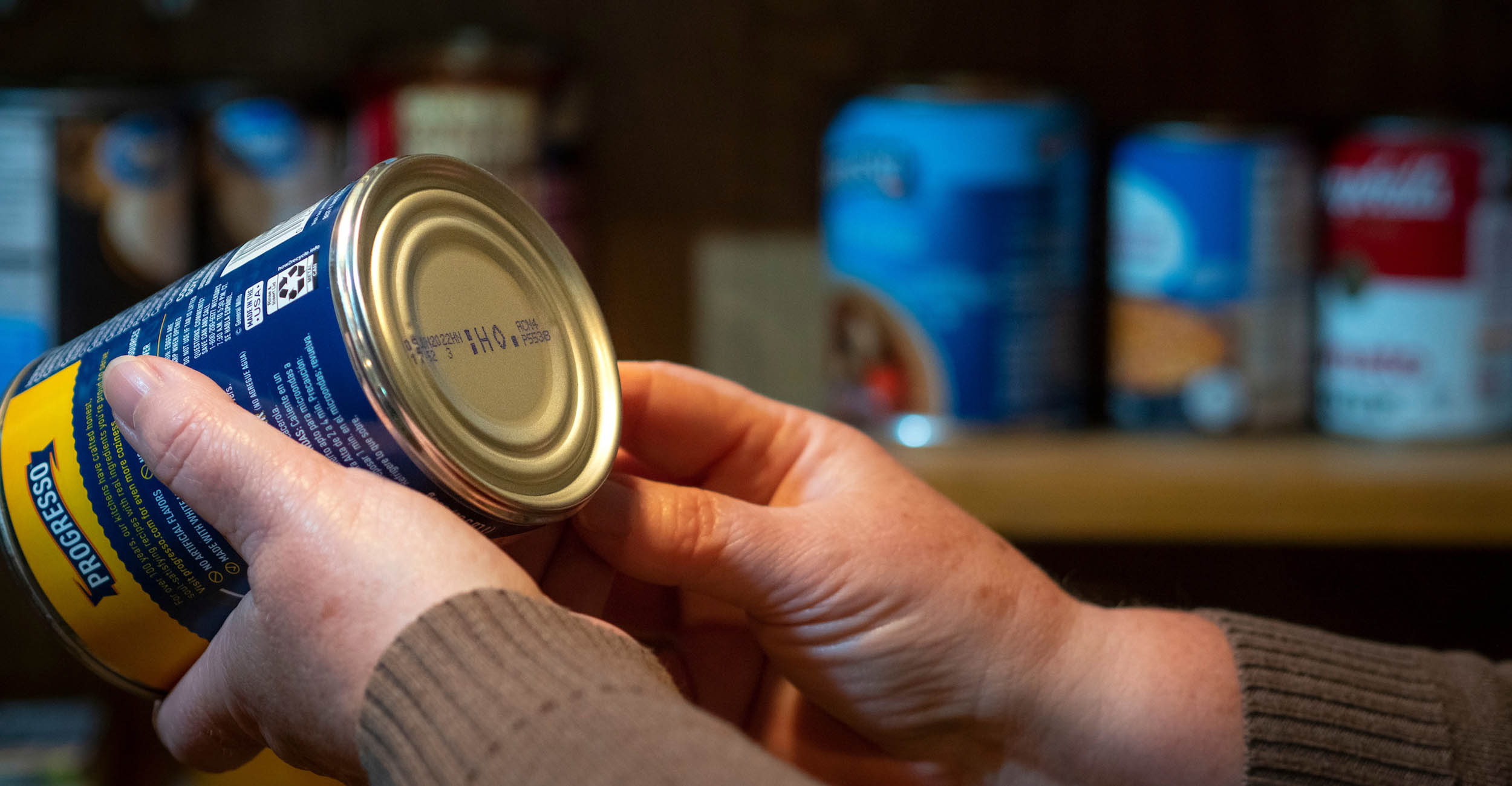 A woman's hands hold a can of food. The can is tipped downward so that the expiration date on the bottom of the can is visible. The woman is wearing long sleeves that are tan in color.