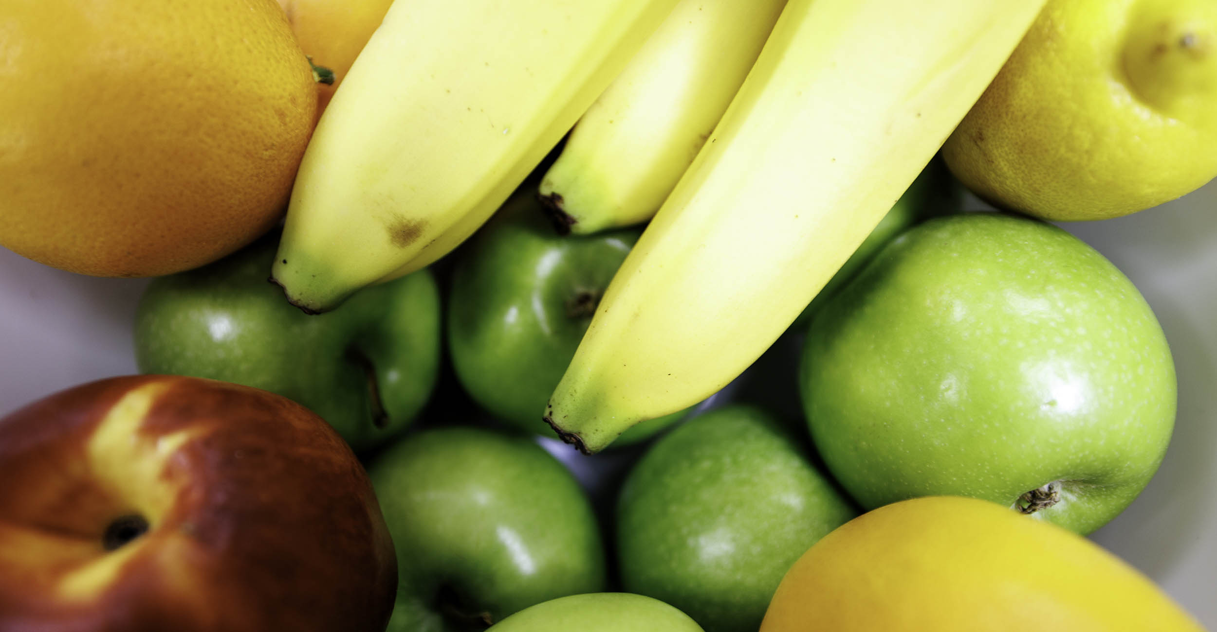 A close-up photo of fresh produce with a white background. The photo includes a red apple, three yellow bananas, an orange, yellow mango and group of green apples.