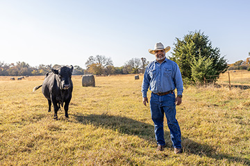Todd Bradford stands in his pasture next to a cow. He wears a cowboy hat, a denim shirt and blue jeans.
