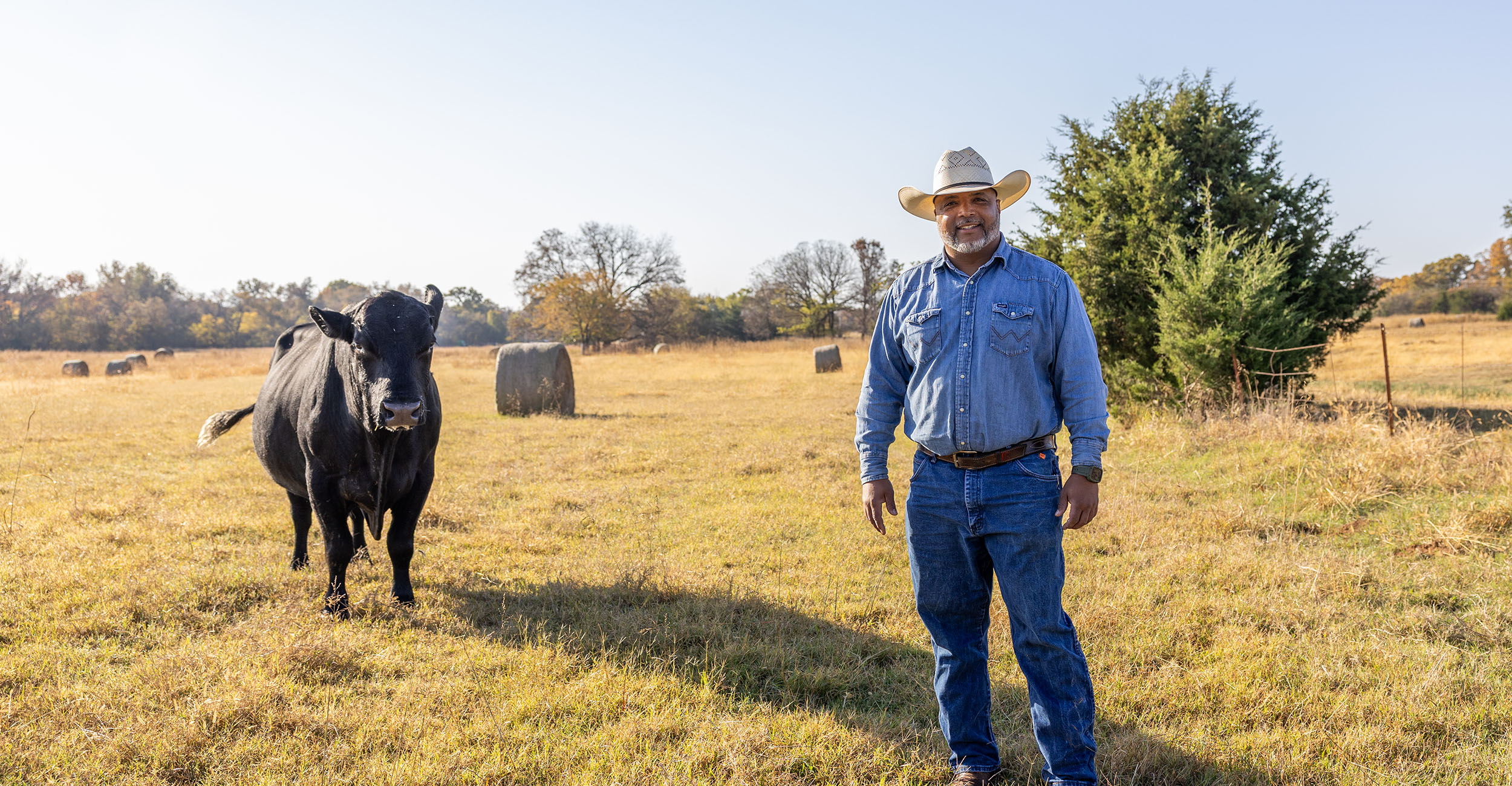 Todd Bradford stands in his pasture next to a cow. He wears a cowboy hat, a denim shirt and blue jeans.