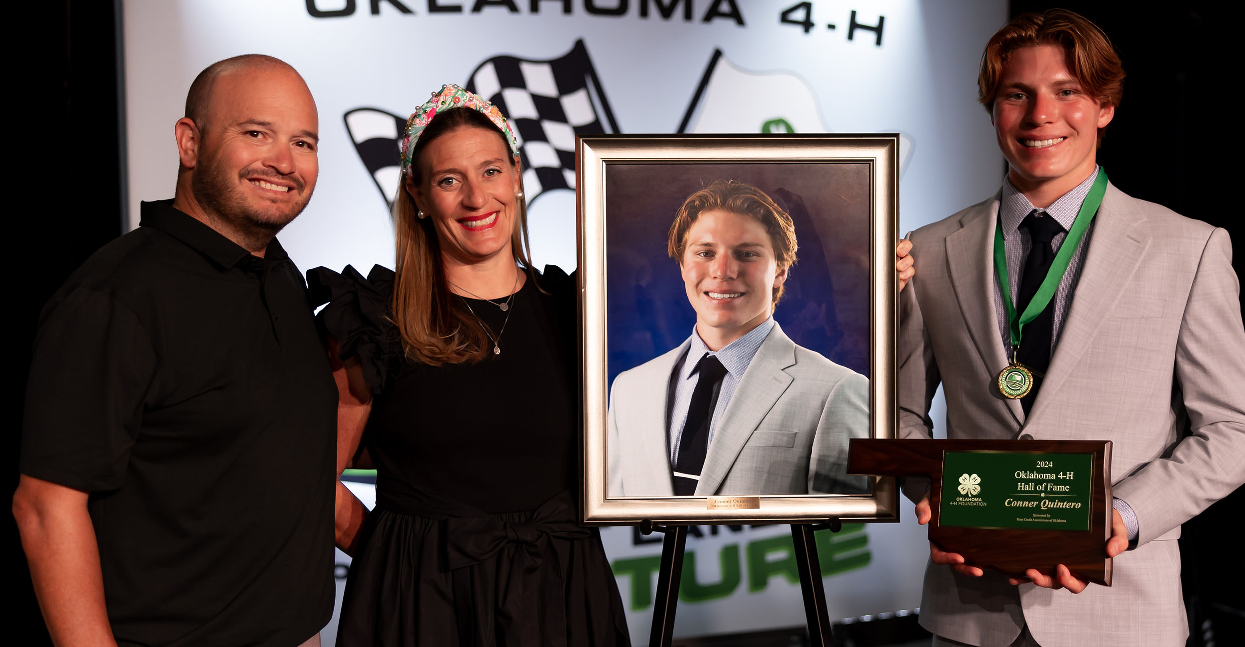 Young man in gray suit on a stage holding an Oklahoma-shaped plaque standing next to his portrait. Also pictured are a man in a black shirt and a woman in a black dress.
