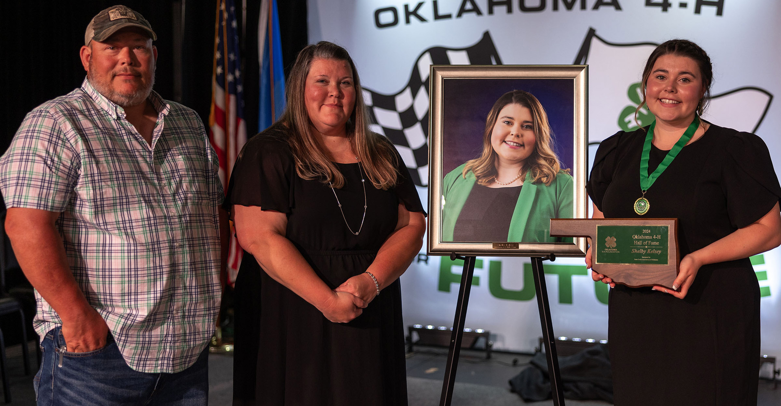 Girl in black dress holding an Oklahoma-shaped plaque and standing next to a large photo of herself. Her parents are also standing next to the photo.