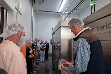 Two people in hairnets examine a packaged cut of beef inside a food processing facility while a group of observers watches nearby.