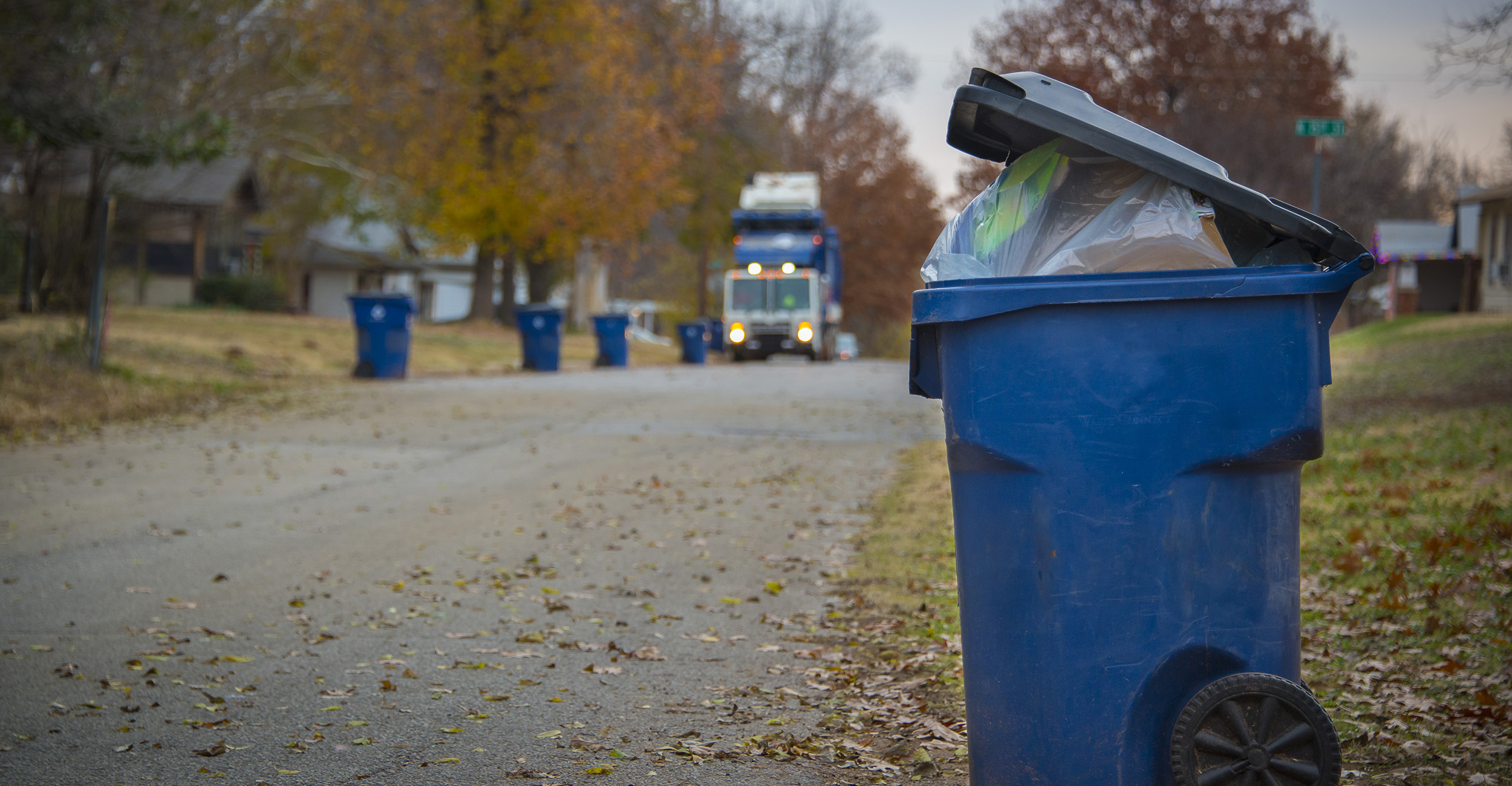 Large blue trash bin stuffed with garbage sits at the edge of a tree-lined street waiting on the trash truck coming down the road.