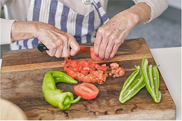 A person holding a chef's knife with a black handle who is slicing a tomato and jalapenos on a wooden cutting board.