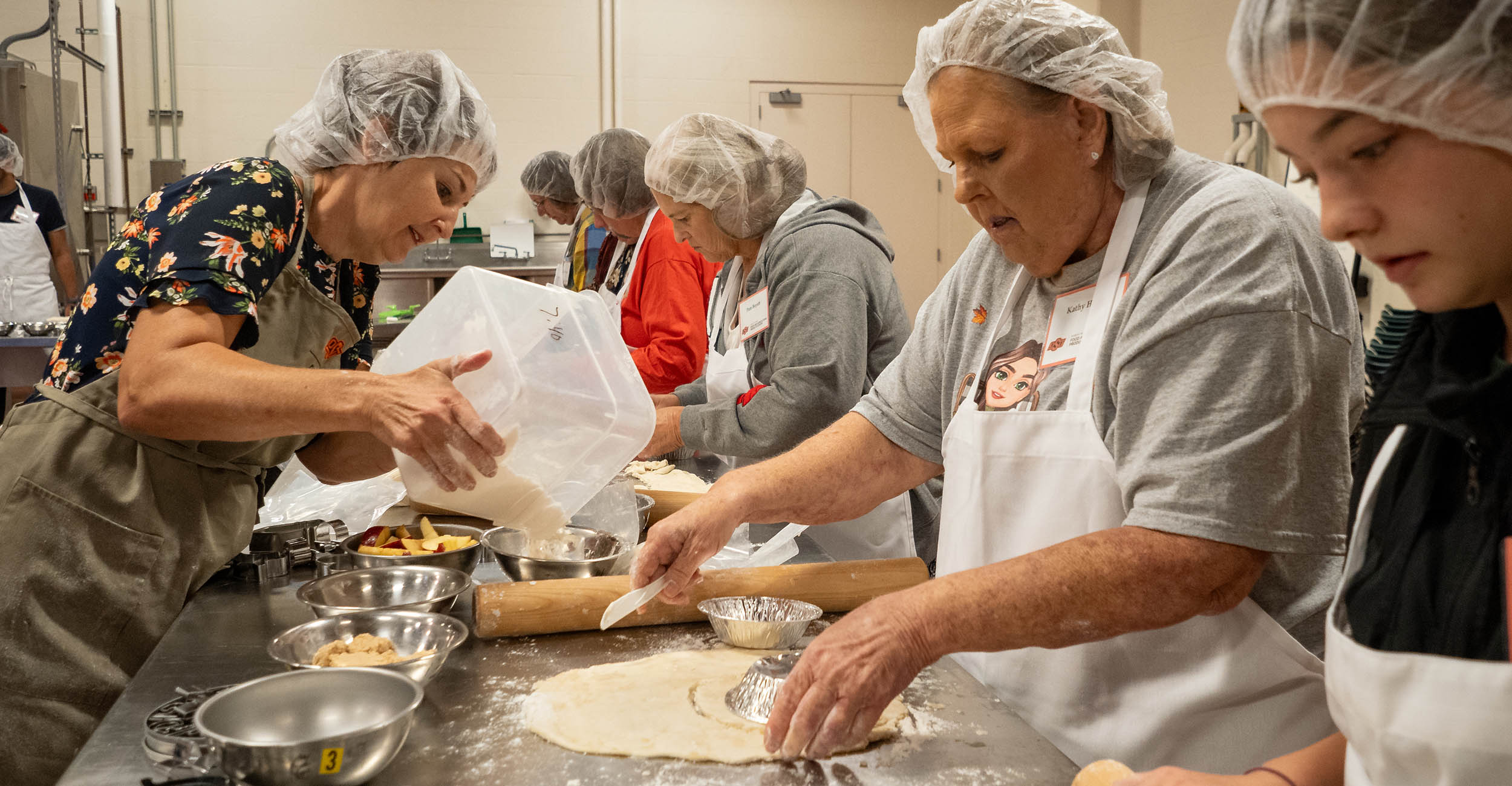 Women of various ages in hair nets and aprons stand alongside a metal baking table preparing pie dough. Baking ingredients, pans, rolling pins and flour are scattered up and down the long table.