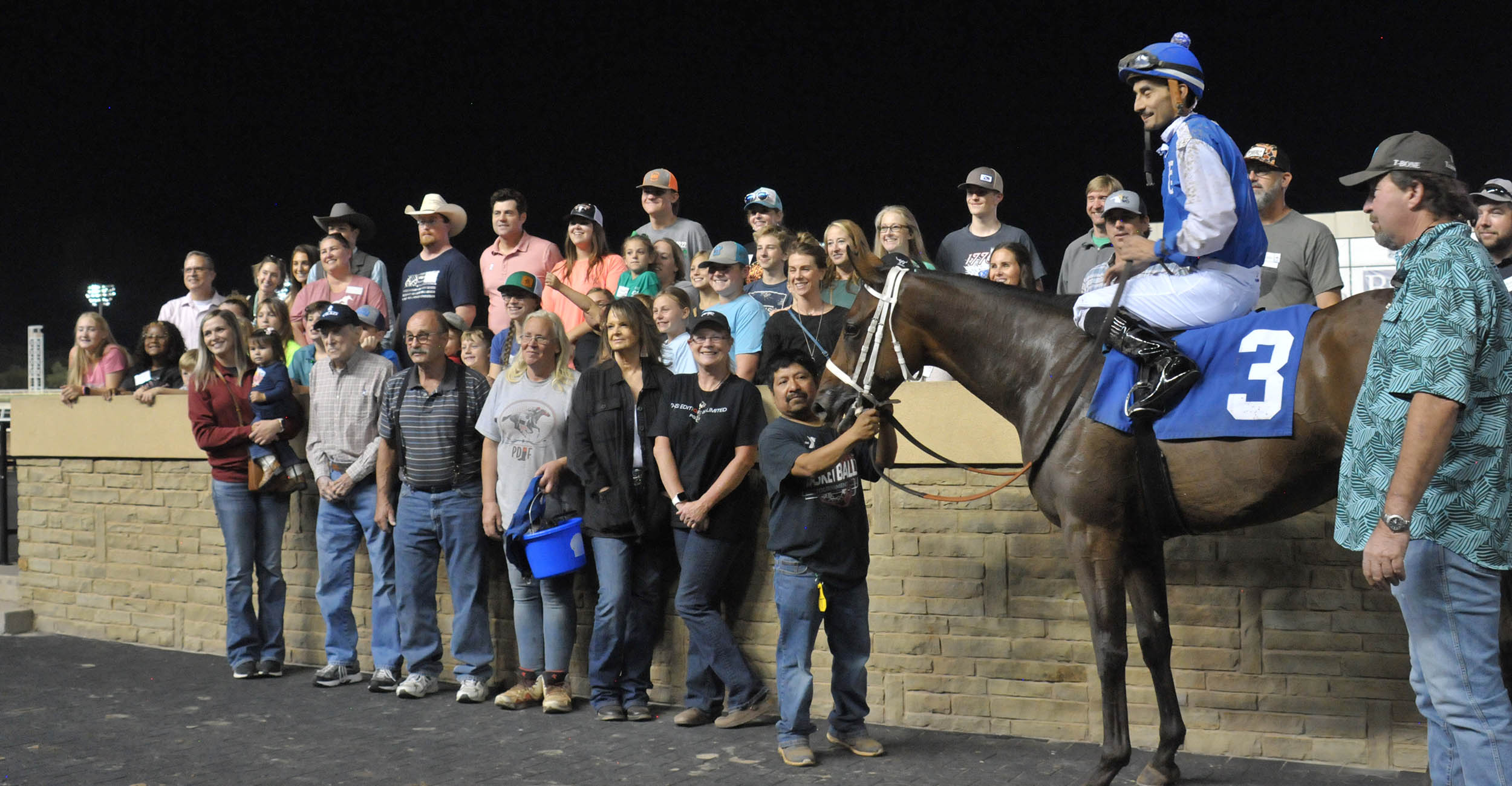 Oklahoma 4-H members, their parents and volunteers stand behind and in front of a tan brick wall that is waist-high alongside a jockey riding blue and white racing silks atop a bay horse with the number 3 on its saddle blanket. The horse's groom is at the horse's head.