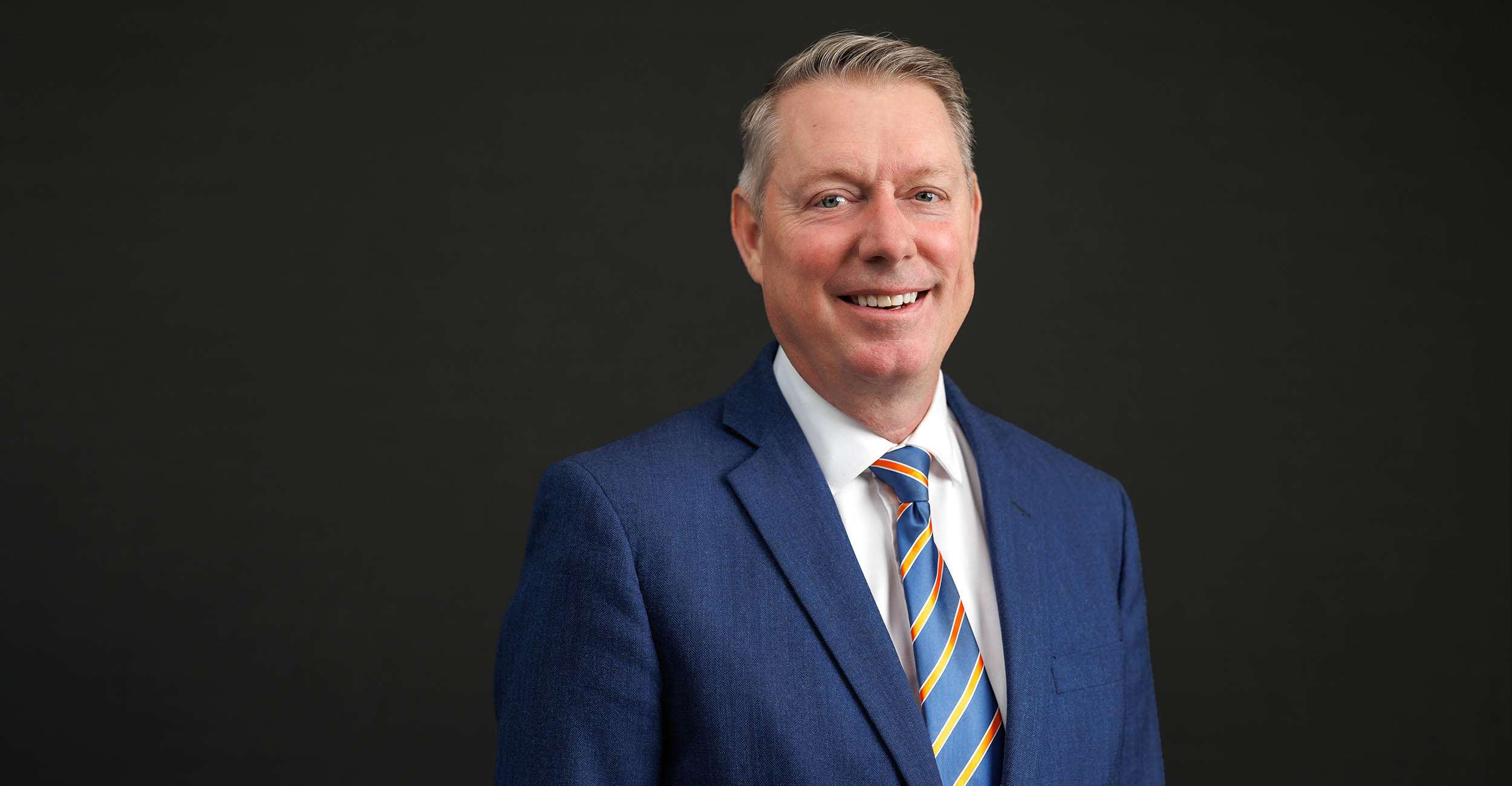 Sleek headshot of Jayson Lusk with a black studio background. He's wearing a navy suit, white dress shirt and blue and orange striped tie..