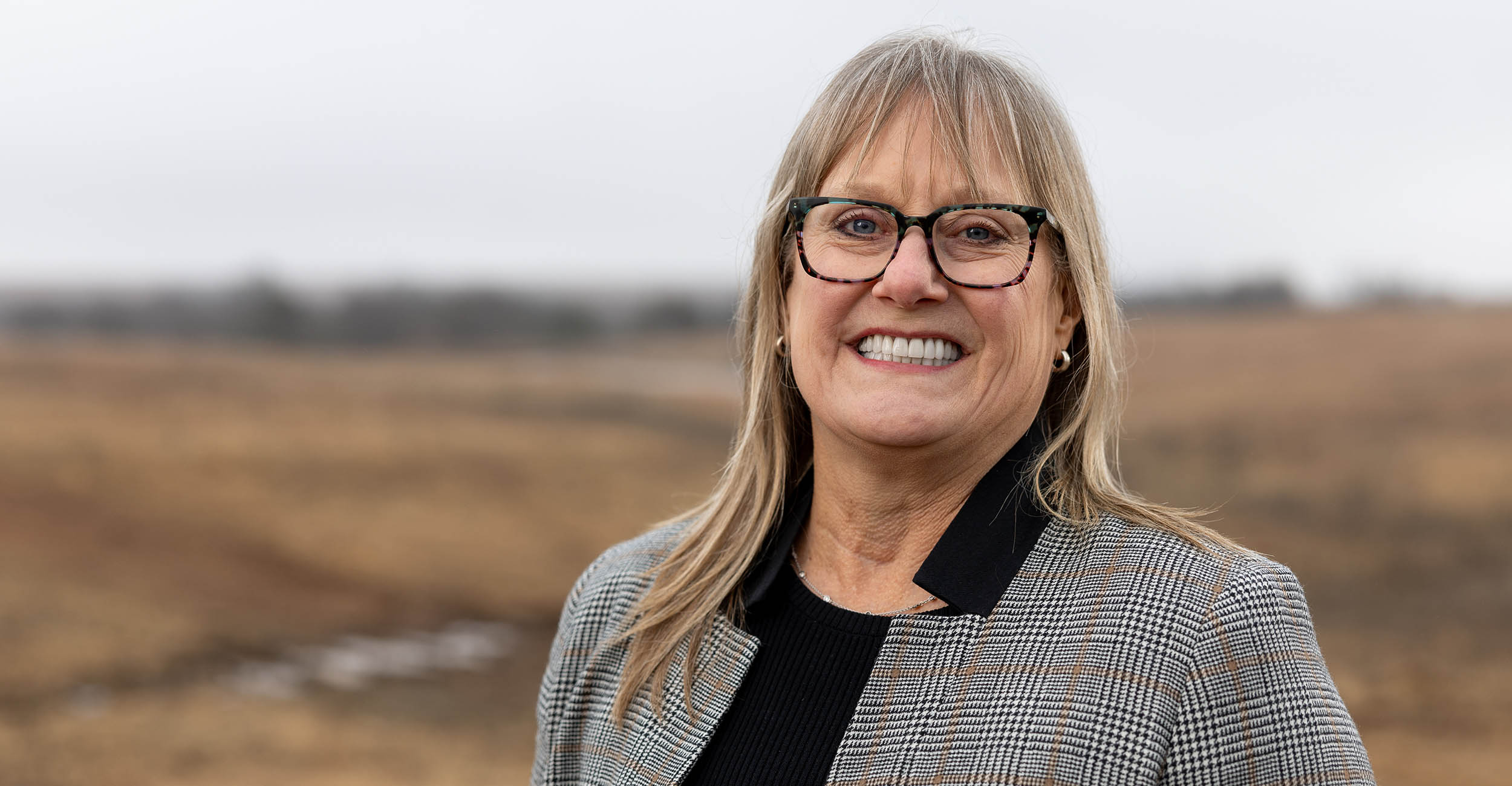 Karen Hickman with her hair down around her shoulders, wearing black glasses. She's wearing a black shirt with a tan, black and white tweed patterned blazer over it, standing in a pasture with brown, dead grass.