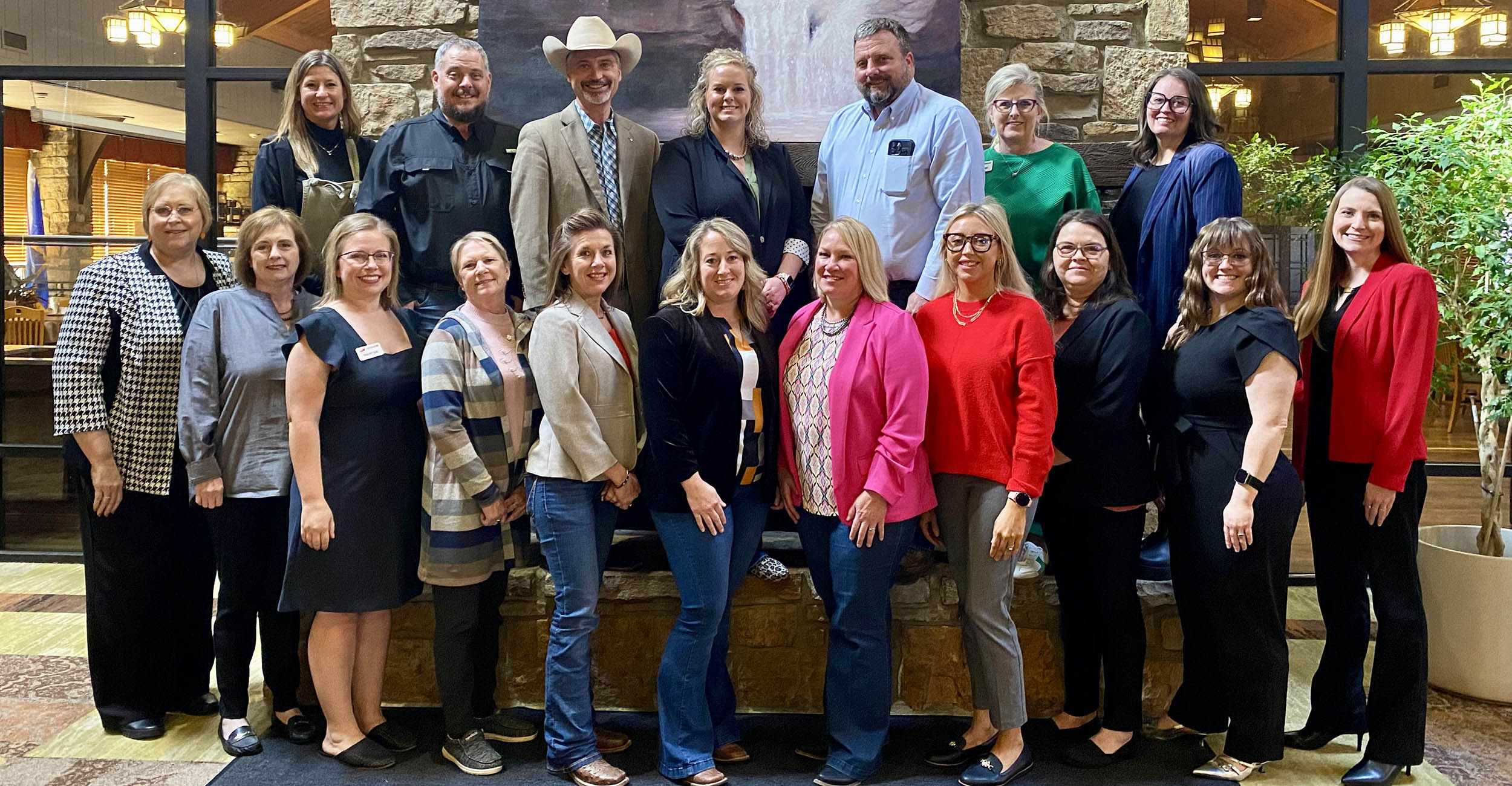 A group of 18 men and women in business casual attire stand in front of a rock fireplace. They stand in two rows. The top row is standing on the seat ledge of the tan rock fireplace.