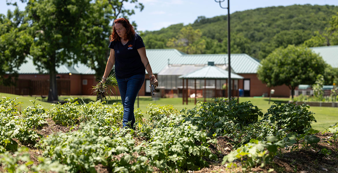 A lady with long, curly red hair wearing a black polo shirt walks through a garden.