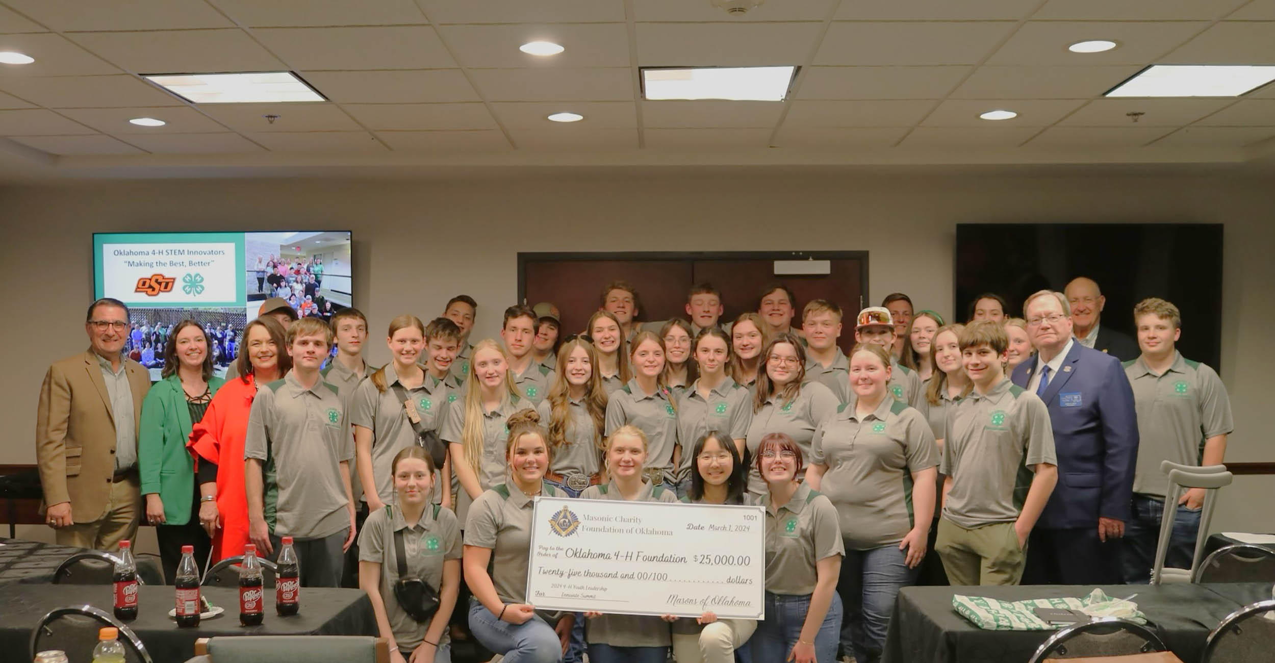 A group of around 35 male and female teenagers stand at the front of a presentation room behind a large print check. The cardboard check represents a $25,000 donation to Oklahoma 4-H from the Oklahoma Masonic Charity Foundation. The teens are wearing tan and green polo shirts and a big screen TV behind the group displays the Oklahoma State University and 4-H logos.