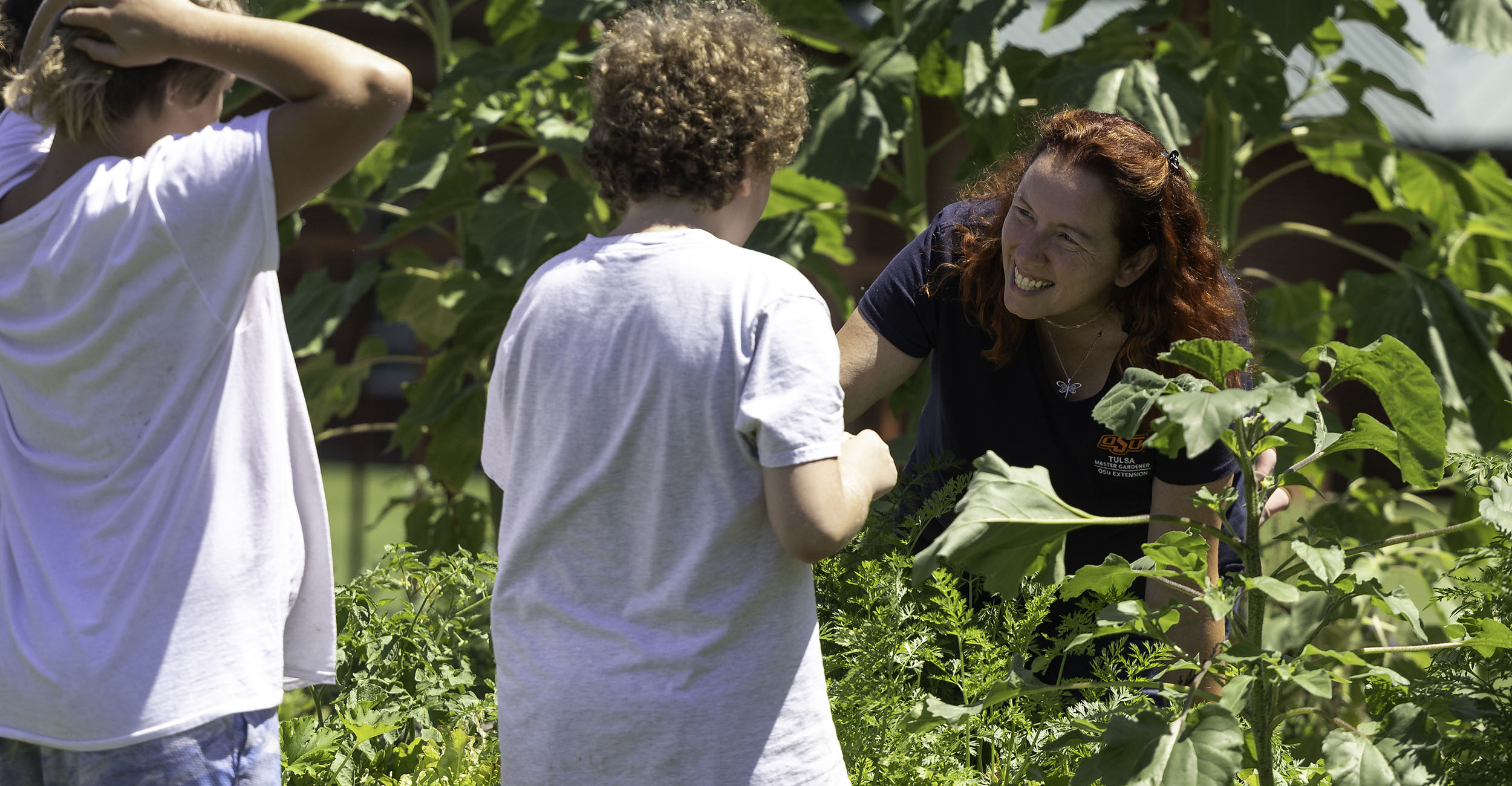 Woman with red hair works in a raised bed garden and teaches two boys various gardening skills.