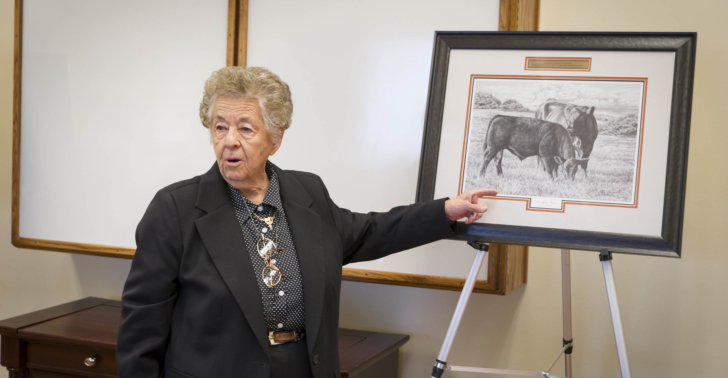 Minnie Lou Bradley stands next to an easel, pointing at the painted picture displayed on it. The picture is of Angus bulls. Minnie Lou is wearing black pants, a black blazer and a black and white polka dot button up collared shirt. She has a pair of glasses hanging around her neck.