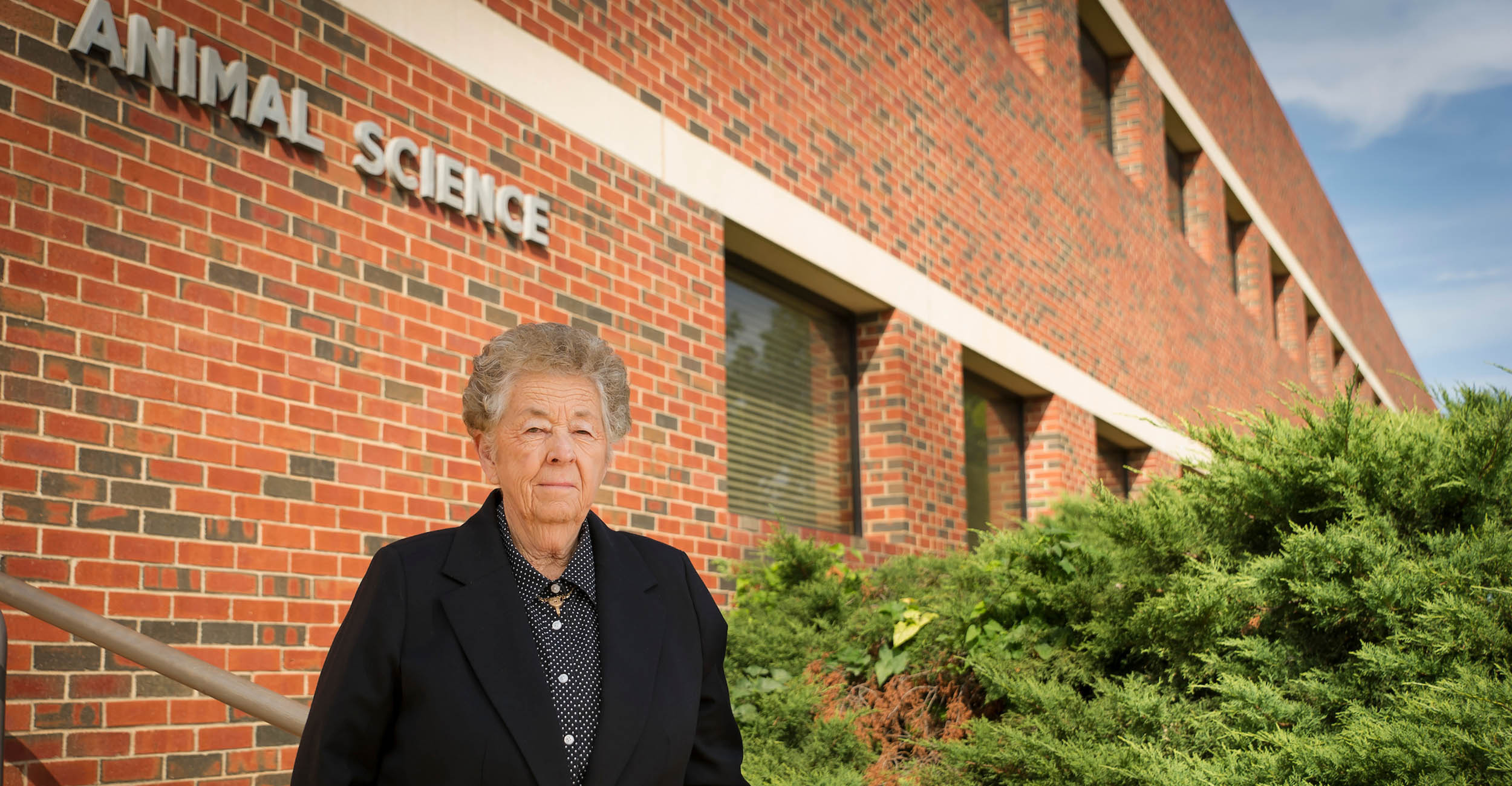 Minnie Lou Bradley stands in front of the main entrance of OSU's Animal Science building of orange brick. The words "Animal Science" are visible on the building behind her head. She is wearing a collared, black and white polka dot shirt with a black blazer over it. There is green shrubbery in front of the building, behind her on the right side.