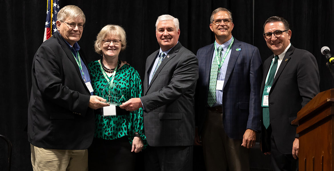 Rodd and Dona stand with three other men to accept their award, which is a clear, glass plaque. Rodd is wearing glasses, khaki pants and a black jacket. Dona is wearing glasses, black pants and a green and black blouse.