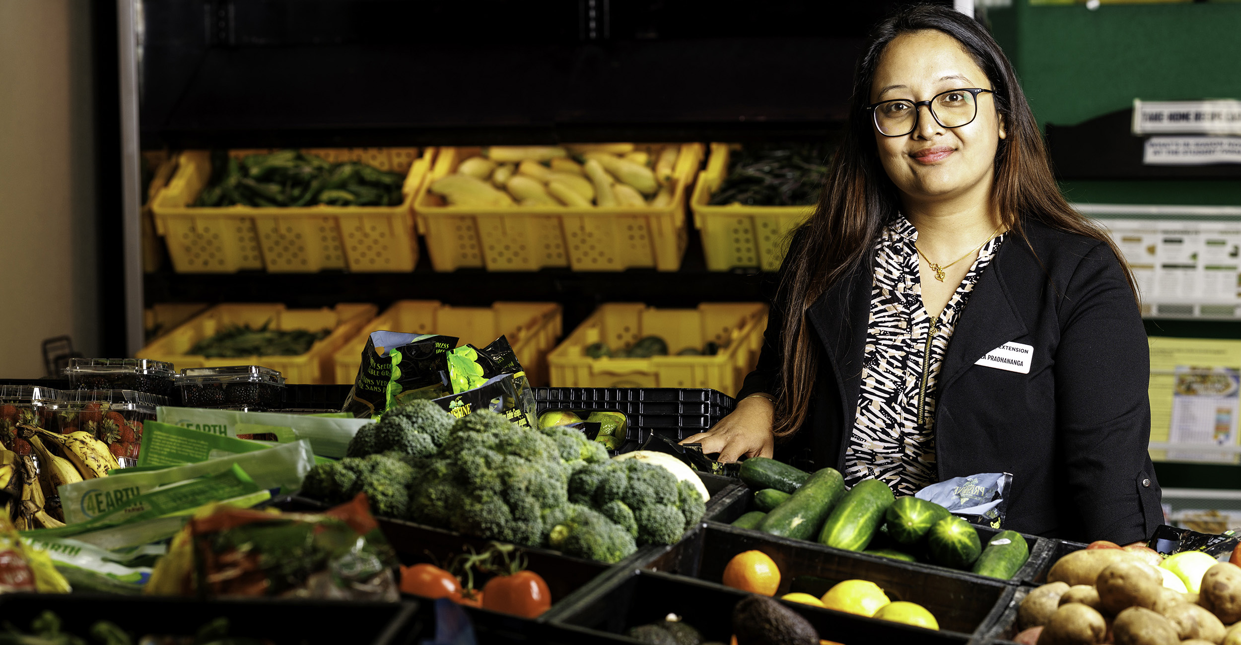 Woman with long, dark hair, wearing glasses and a black sweater, stands behind several crates of fresh vegetables.