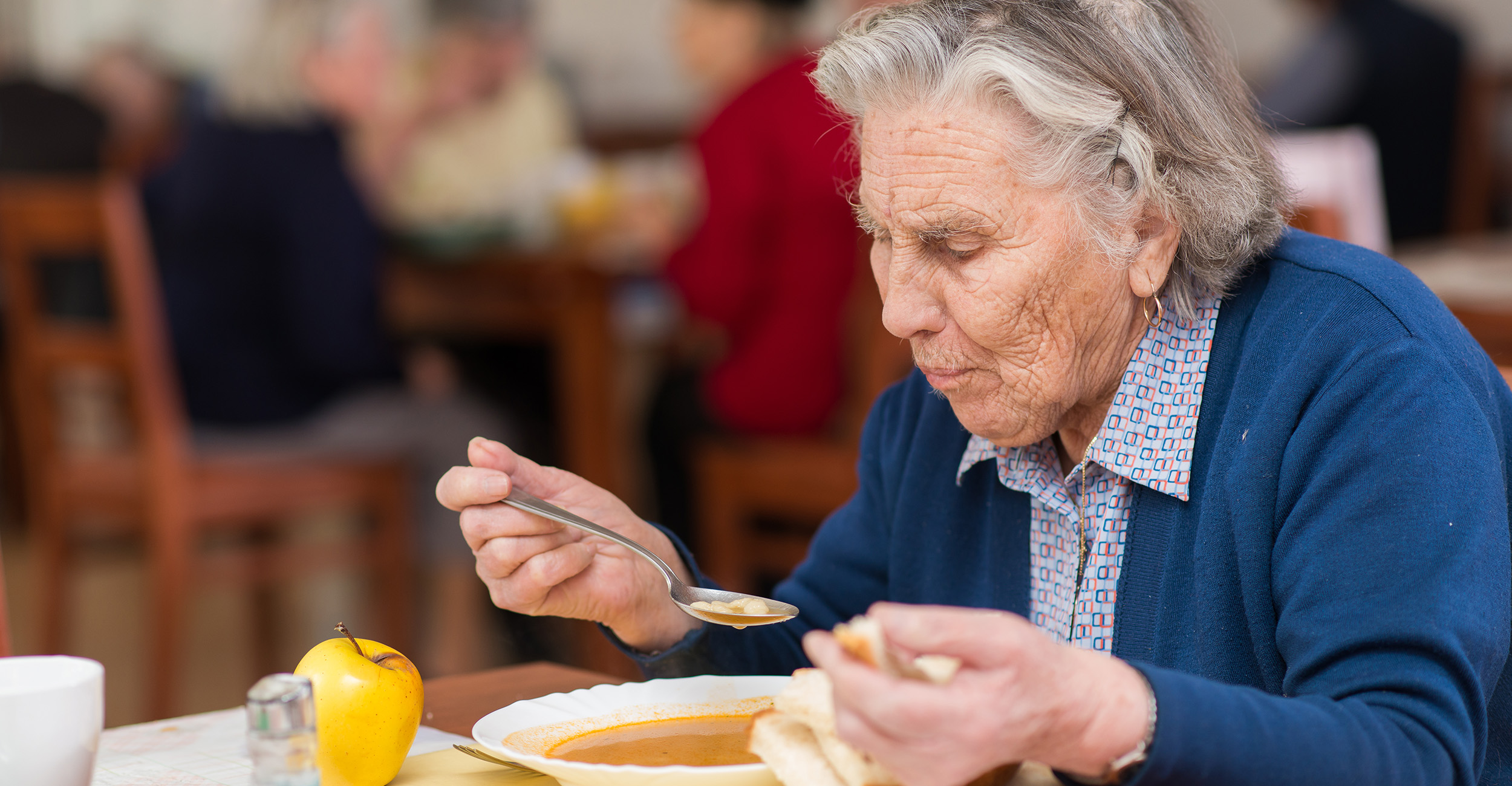 Woman with gray hair and wearing a blue sweater is eating a bowl of soup and a slice of bread.