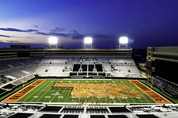 Aerial view of Boone Pickens Stadium at dusk, with the field illuminated and a large crowd of people gathered on the field in the shape of an Oklahoma State University logo.