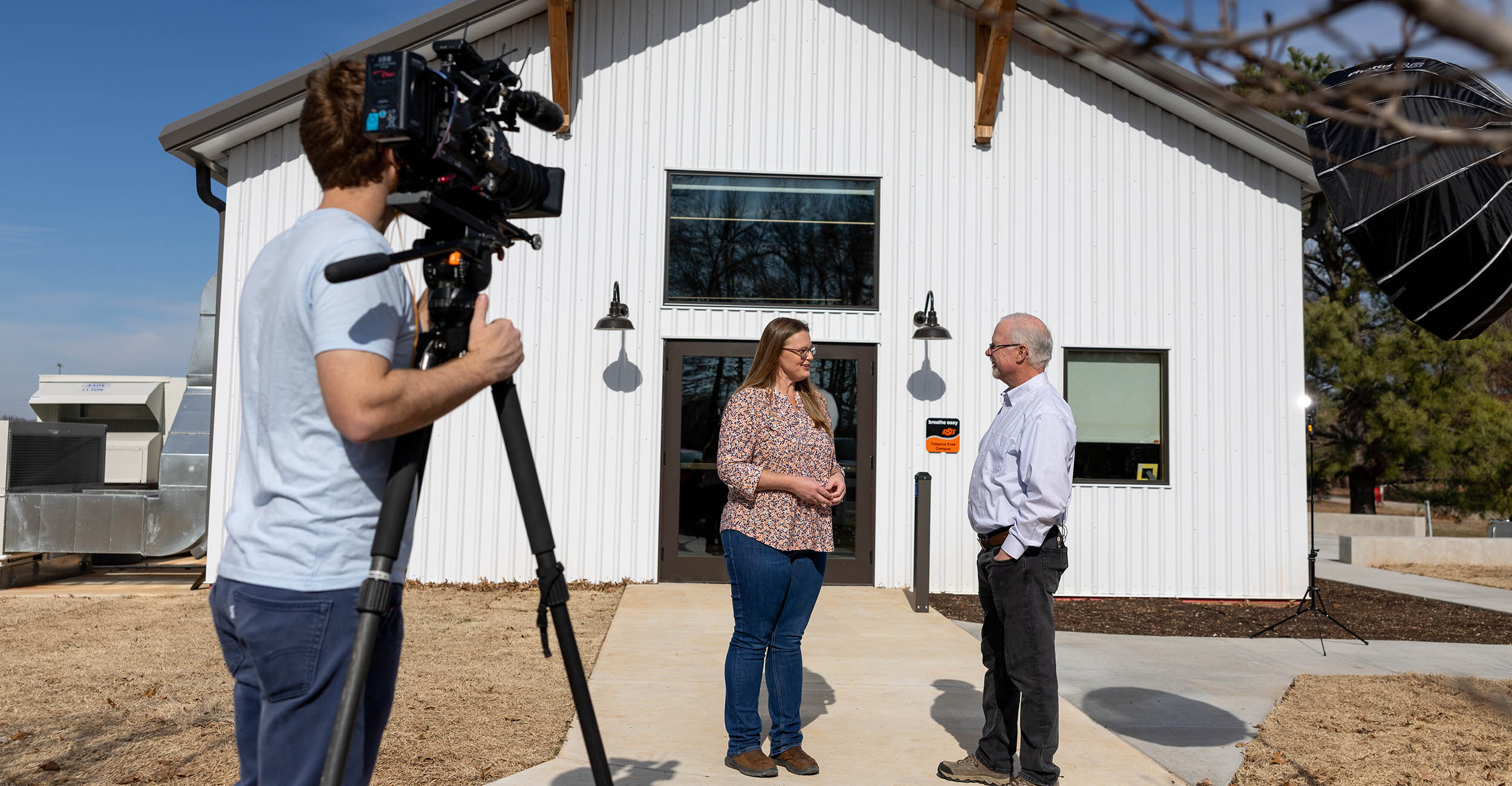 a man in blue jeans and blue T-shirt stands with a video camera in the left side of the photo videoing a woman in a flower print shirt and blue jeans as she interviews a man in a blue shirt and blue jeans to her right. The man and woman in conversation are standing on a sidewalk in front of the entrance to the new Horticulture Education Center building that is a rustic barn design with white siding at The Botanic Garden at Oklahoma State University.
