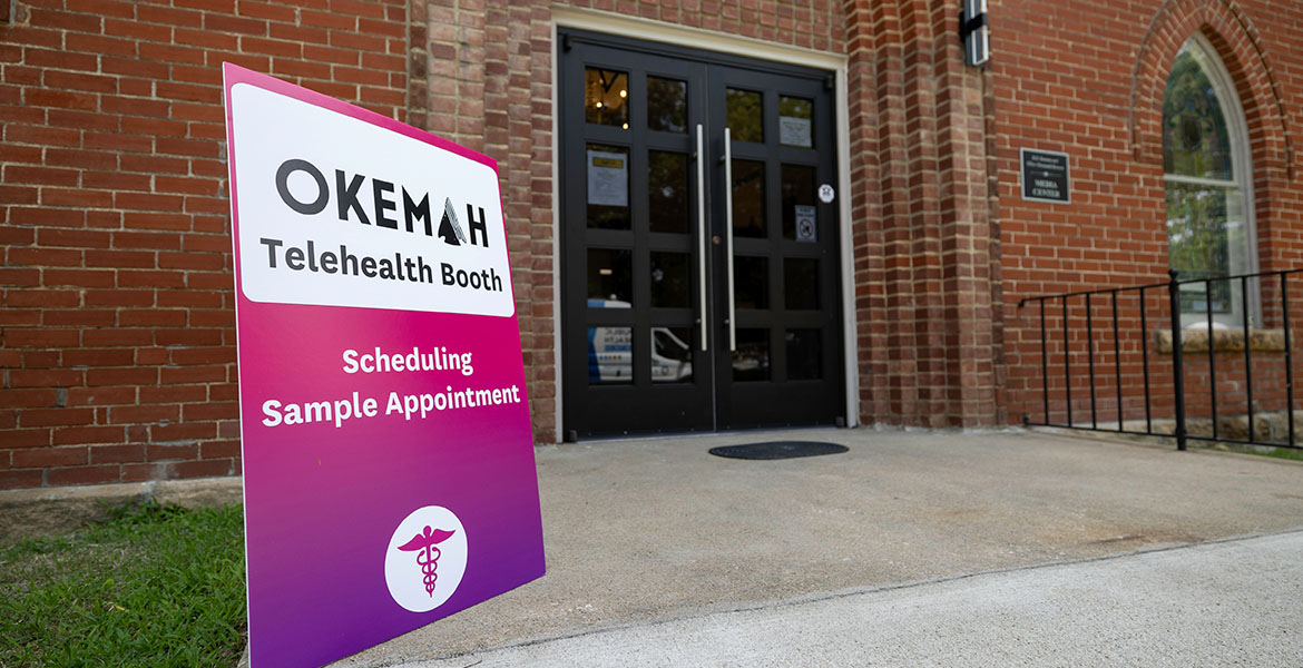 A bright purple and pink sign sits at the front entrance of the Okemah Library Media Center, which is an old brick church building. The sign says "Okemah Telehealth Booth Scheduling Sample Appointment."