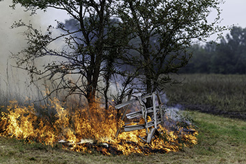 A fire burns through dry grass and vegetation near a fence and tree, with flames and smoke filling the background.