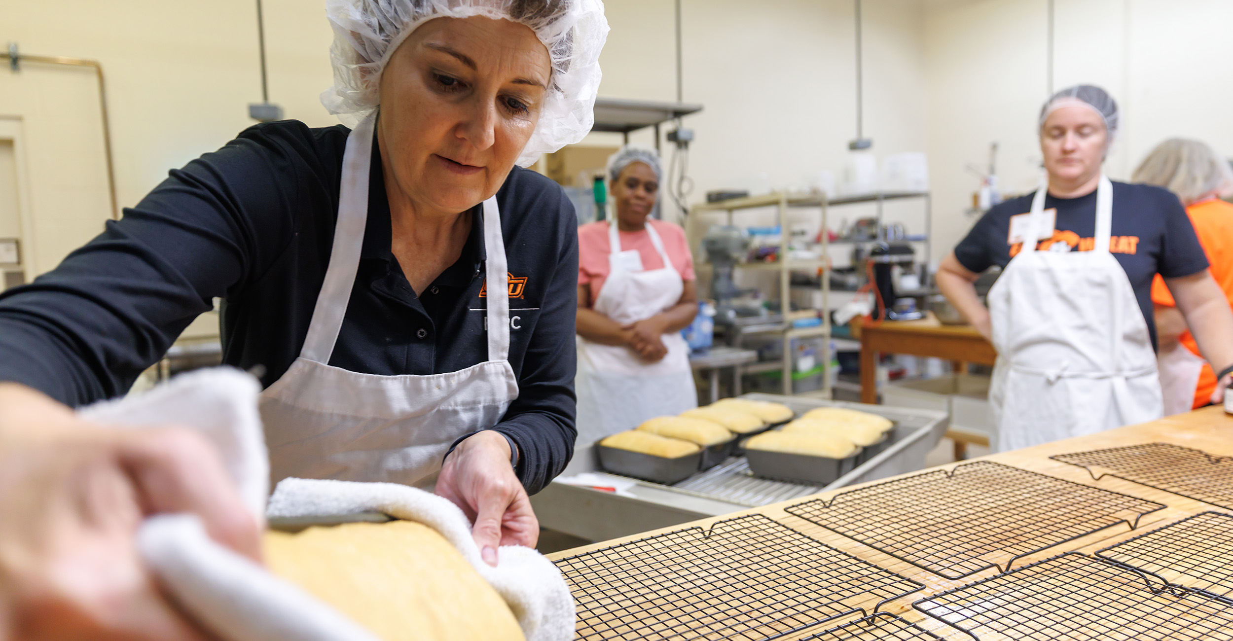 Woman wearing a white apron, black shirt and white hairnet, places a pan of sourdough bread on a cooling rack..