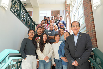 A group of university faculty, staff and students poses together on a staircase inside a sunlit brick building.