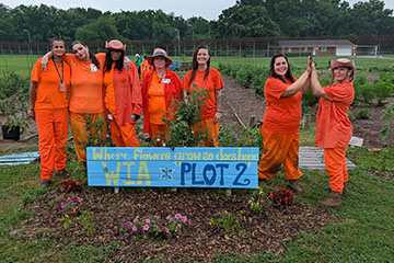 Seven women in orange prison uniforms pose in a vegetable garden, with two of the women high-fiving beside a colorful sign reading “Where flowers grow, so does hope” and “WIA Plot 2.”