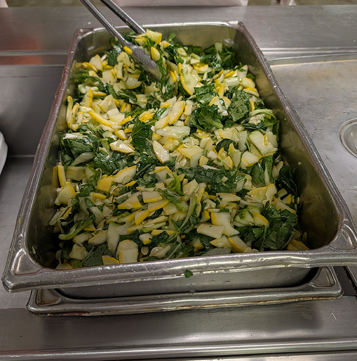 A pan of fresh, chopped vegetables and leafy greens is prepared for service on a stainless-steel food-service tray.