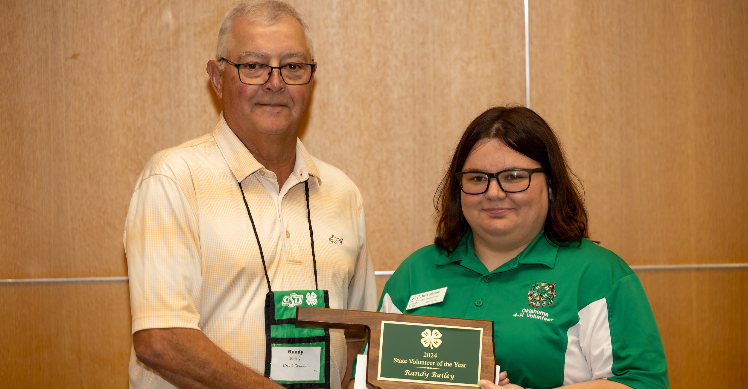 Man wearing collared shirt and glasses is presented an Oklahoma-shaped plaque from a woman wearing a green and white shirt and glasses.