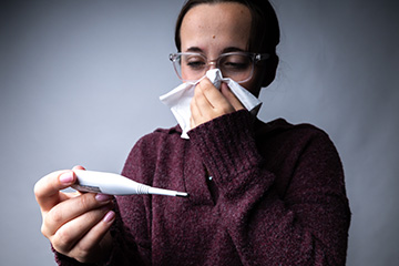 A woman wearing glasses holds a thermometer and uses a tissue to cover her nose, appearing to have cold or flu symptoms.