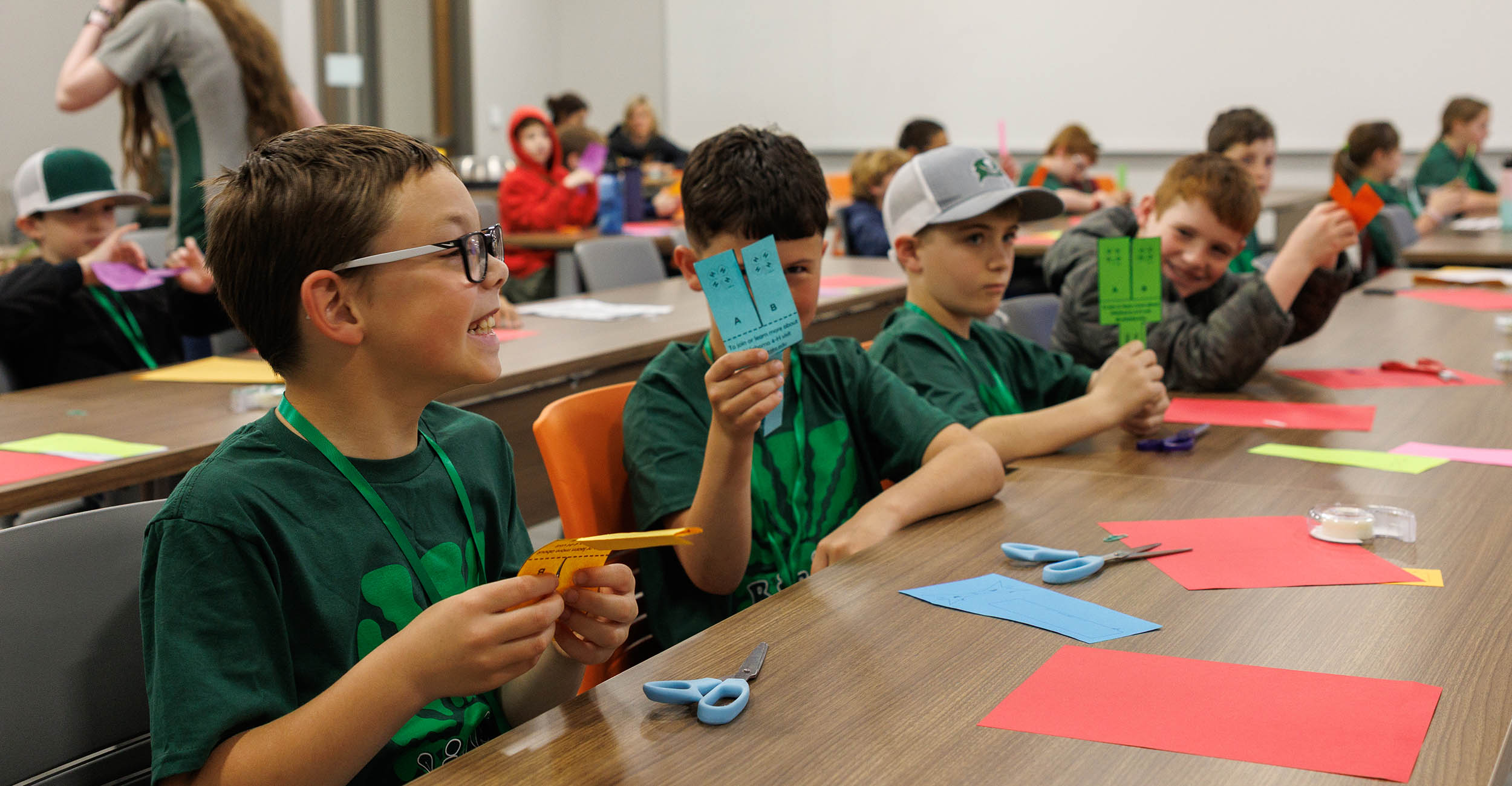Young grade school boys in green T-shirts sit in a row at a long rectangular table holding construction paper for a craft project.