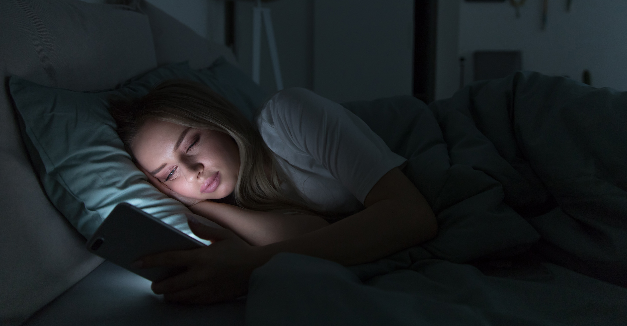 Teenage girl with long, blonde hair, looking at her smartphone while laying on her side in bed.