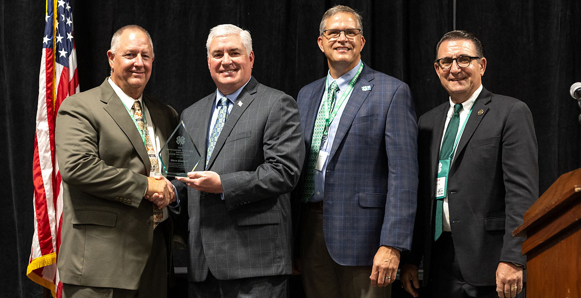 Steve Bomhoff with white hair and wearing a suit holds his clear, glass plaque while shaking the hand of a man wearing a brown suit. Two other men in blue and black suits pose with him in the photo.