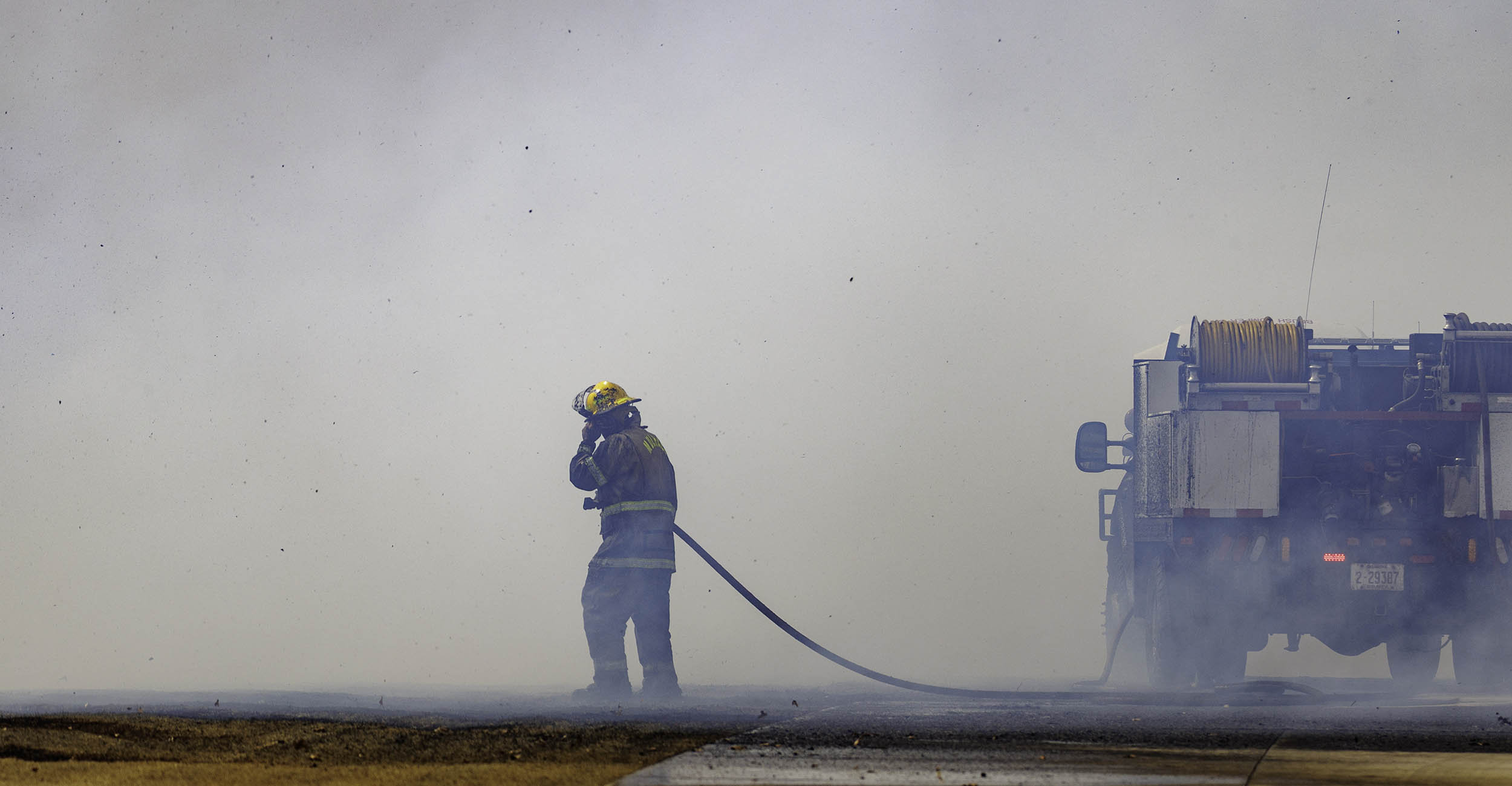 A firefighter in his helmet and yellow and brown bunker gear stands beside a fire truck holding a water hose. He's surrounded by thick smoke and he's rubbing his face.