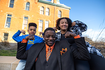 An adult man and two children sit outside in front of a historic brick building, smiling and flexing their muscles in a playful pose. The adult wears a black suit with an orange shirt and tie, while the children stand behind in casual clothing.