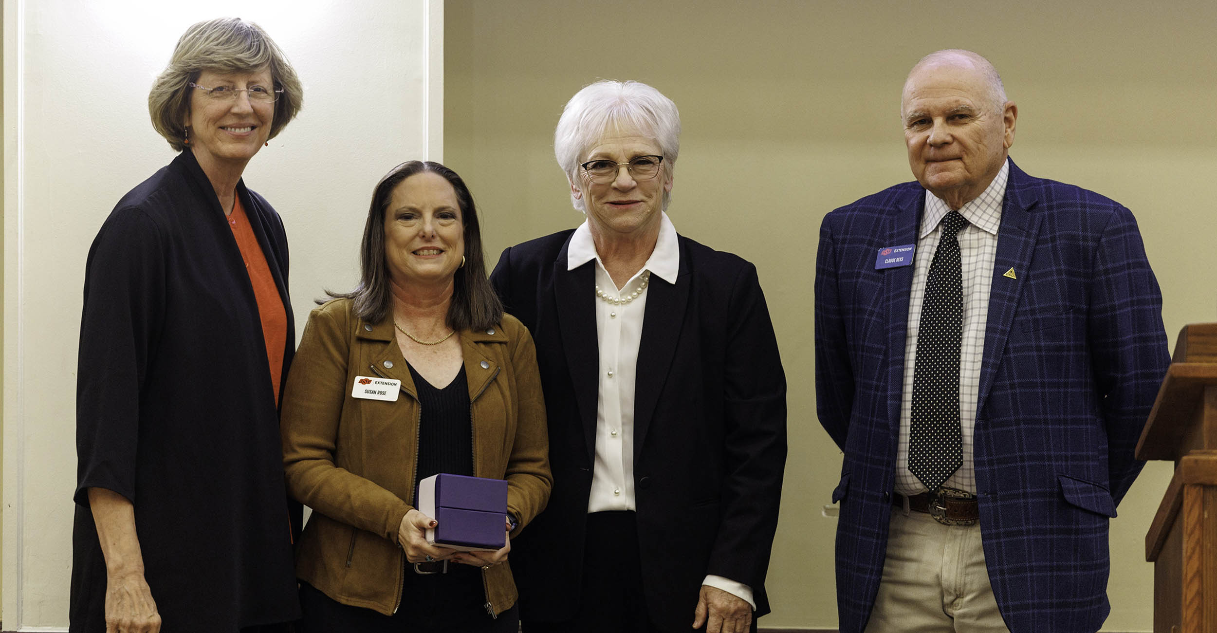 Three women stand with one man on the far right. They are wearing business casual clothes and posing for the camera. The second woman on the left is holding a purple box.