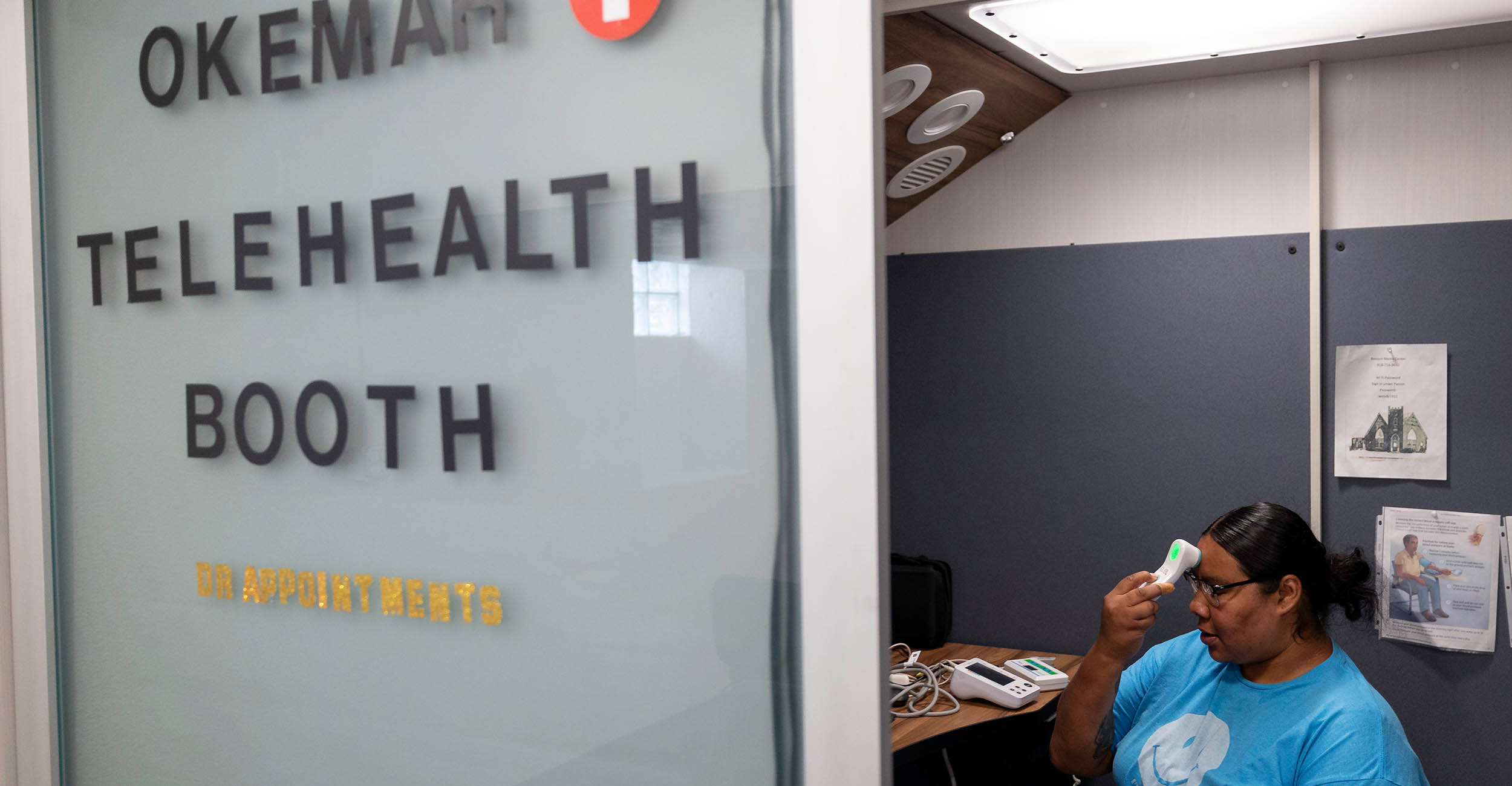 A lady sits in a chair holding a thermometer to her forehead, checking her temperature. She is sitting in a small room in front of a computer. The booth door is labeled Okemah Telehealth Booth.
