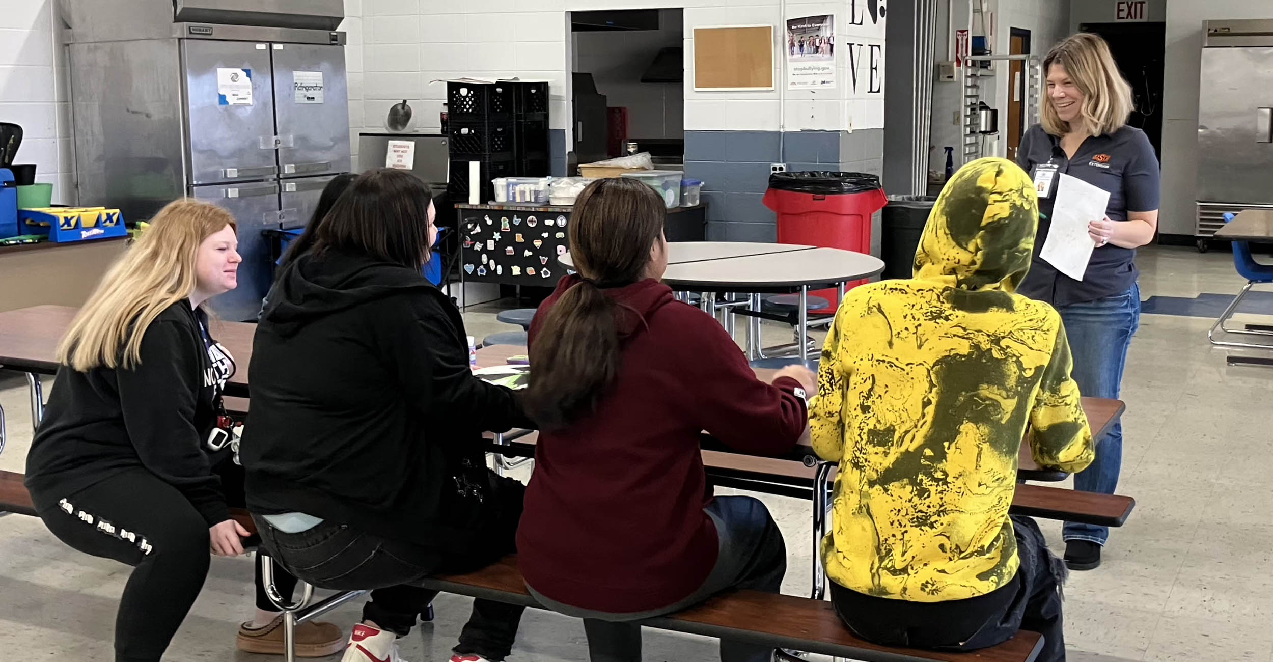 Jamie Foster, wearing a gray OSU polo shirt, jeans and black shoes, stands in a cafeteria, holding a sheet of paper and talking to a group of four high school students, male and female, who are sitting at a cafeteria table listening to her. The students are wearing sweatshirts, jeans, sweatpants and sneakers.