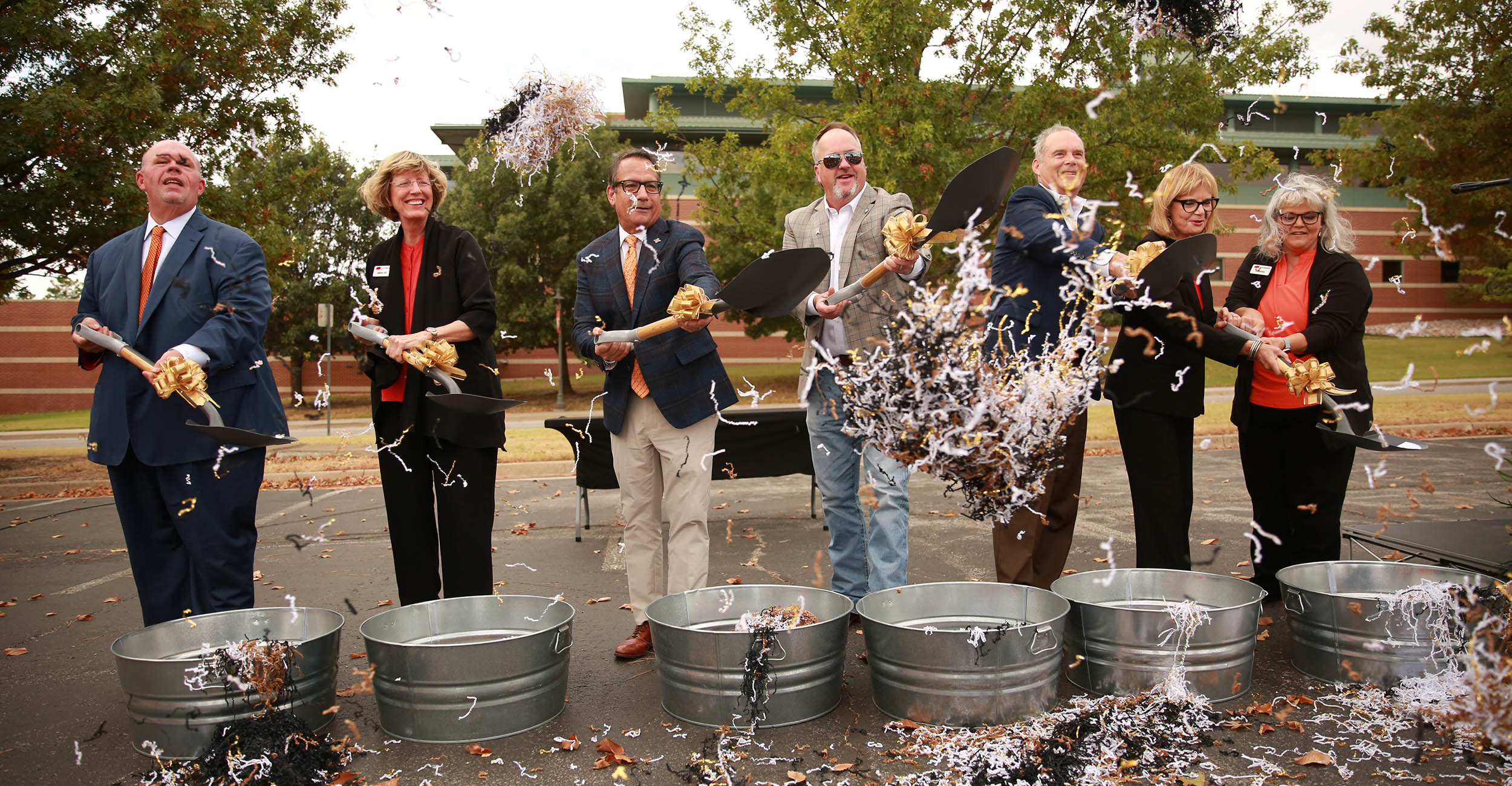 A group of seven men and women wearing black, orange and tan attire scoop gold, black and white confetti out of metal tubs with small shovels. Tree branches and a tan and orange brick building are behind them.