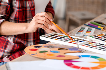 A person in a red plaid shirt uses a paintbrush to mix watercolor paint from a palette, with watercolor paints, a wooden palette and a color wheel visible on a table.