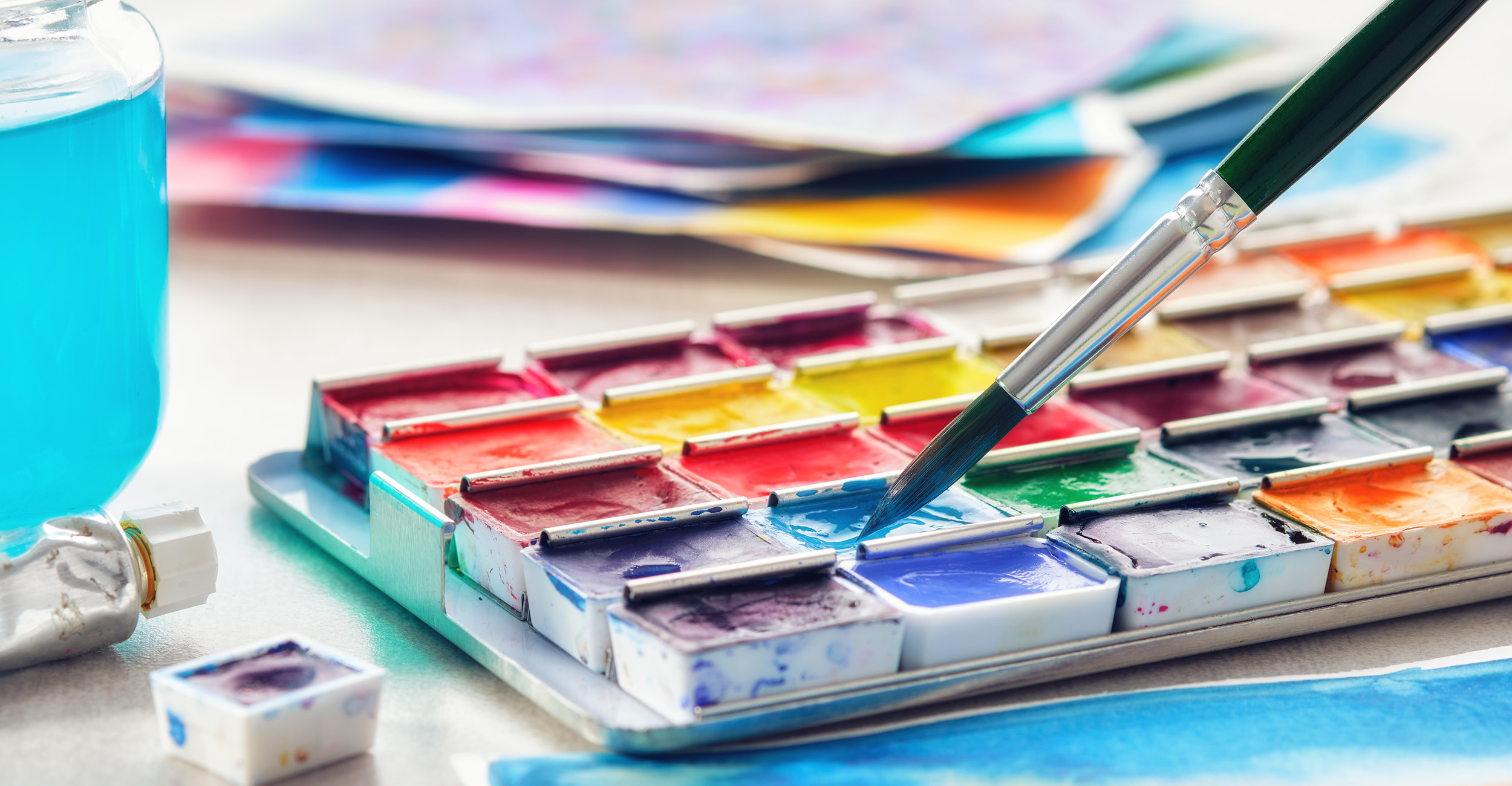 A watercolor paint set with multiple colors sits on a white table with a paintbrush is dipped into one of the colors.