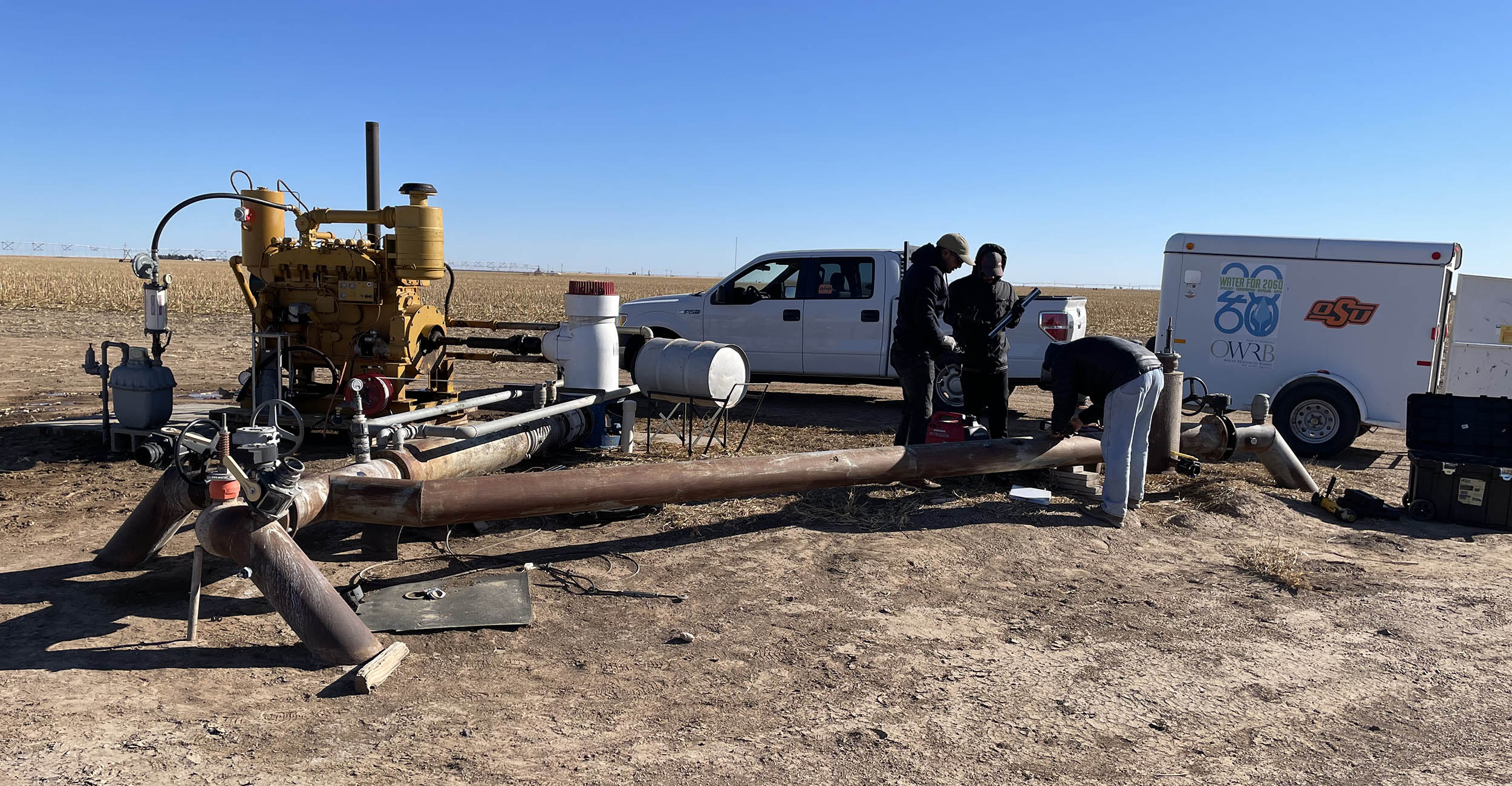 Three men bend over a large pipe that sits above ground at the edge of a field where corn is harvested. A pump system and quad cab pickup truck hooked to a small equipment trailer are also in the photo. The men, in coats and jeans, are testing the water pump.