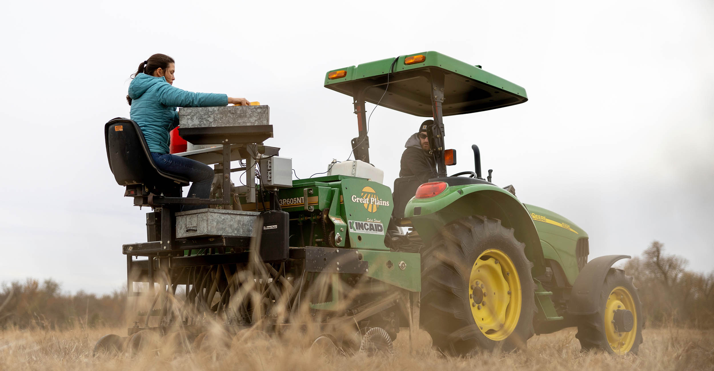 a man drives a small John Deere tractor as a woman in a blue coat sits on a small field planter monitoring the machine as it plants wheat seed