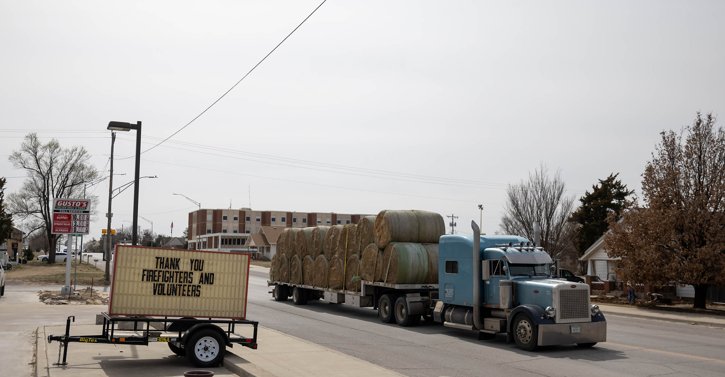 A light blue semi-tractor trailer hauling a load of large round hay bales through a main street of the small town of Shattuck, Oklahoma. A sign parked alongside the street says "Thank You Firefighters and Volunteers."