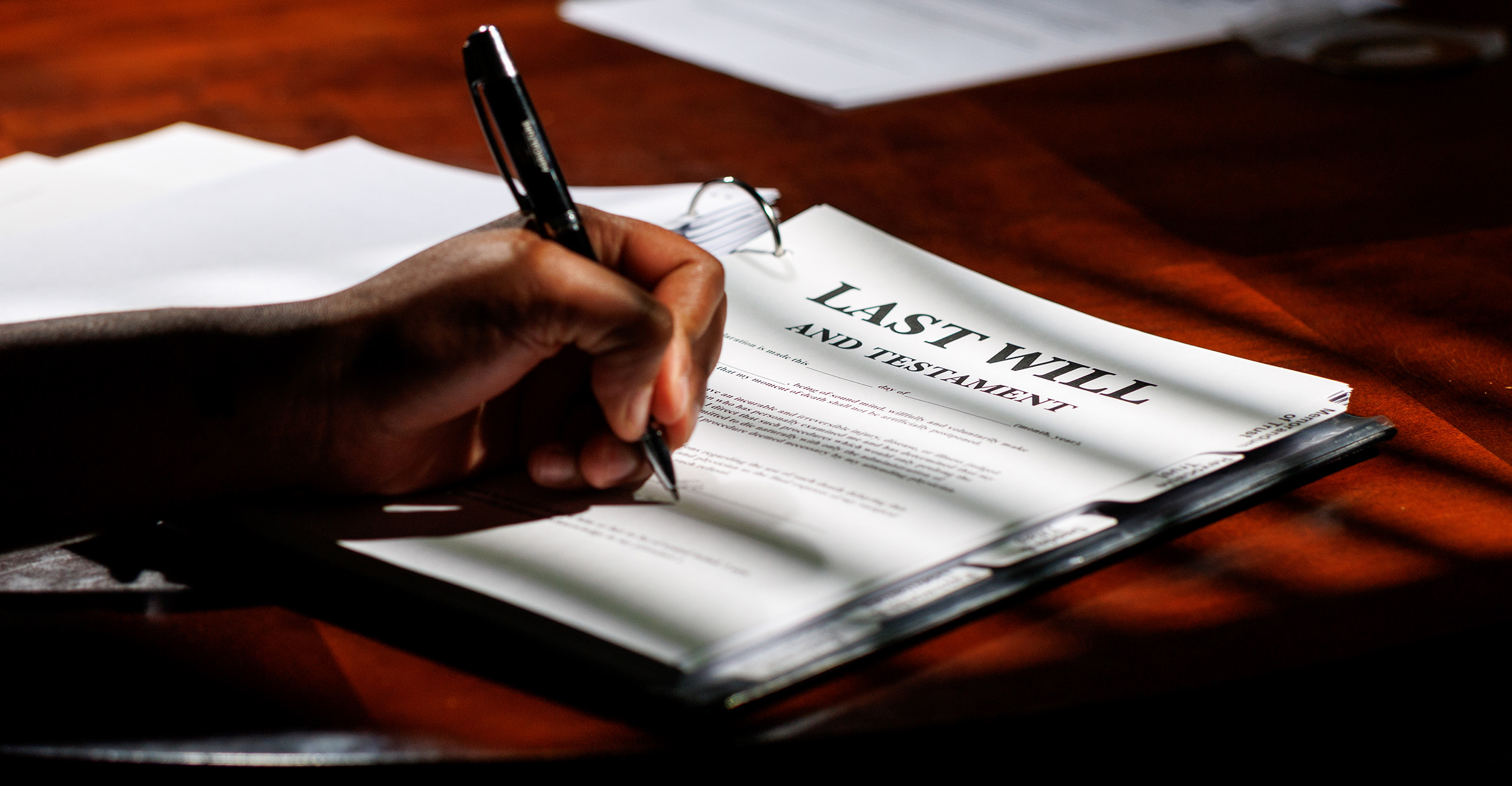 A three-ring binder on a desk containing a will and trust paperwork with a hand holding a pen signing the paperwork.