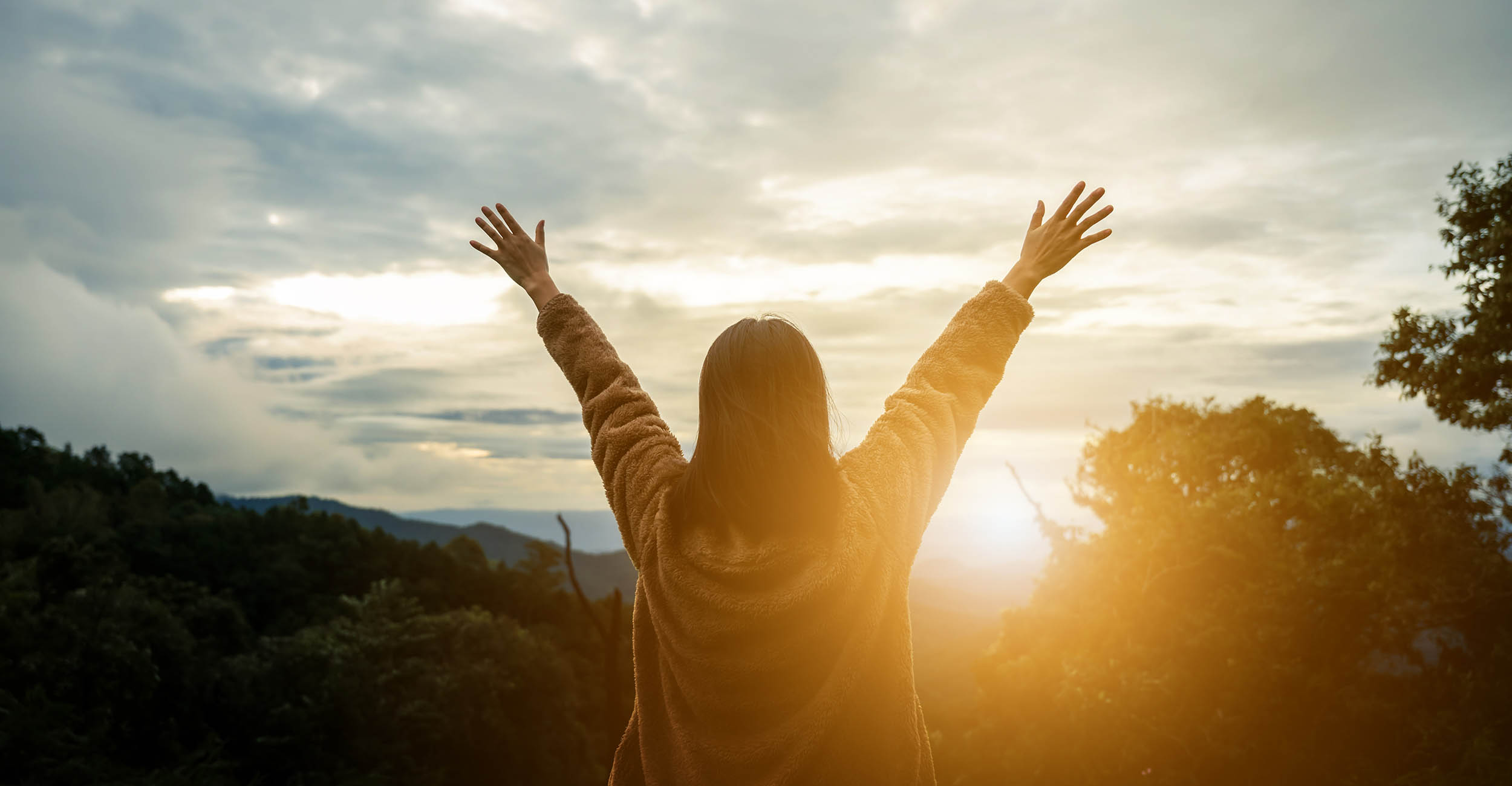 A young woman with her back to the camera stretching her arms and fingers up into the sky at a sunrise or sunset. She has brown shoulder-length hair and is wearing a brown, fuzzy coat.