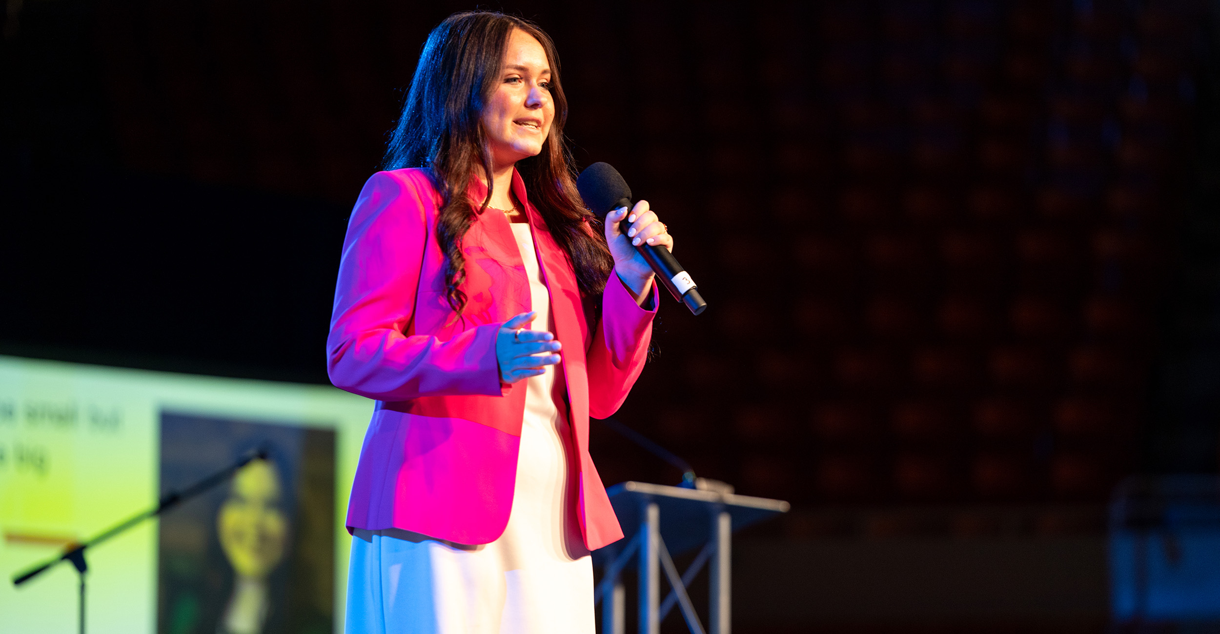 Young woman in pink blazer standing on stage with a microphone.