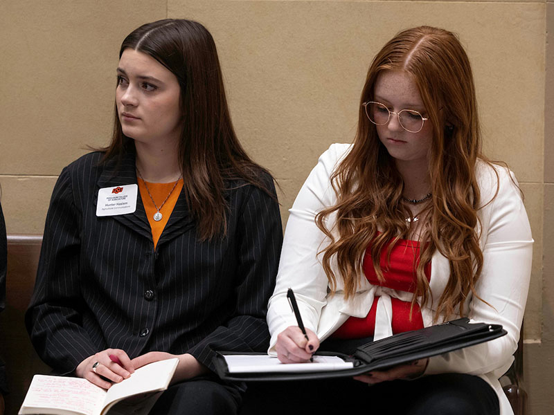 Two women are sitting in chairs against the wall listening and taking notes.